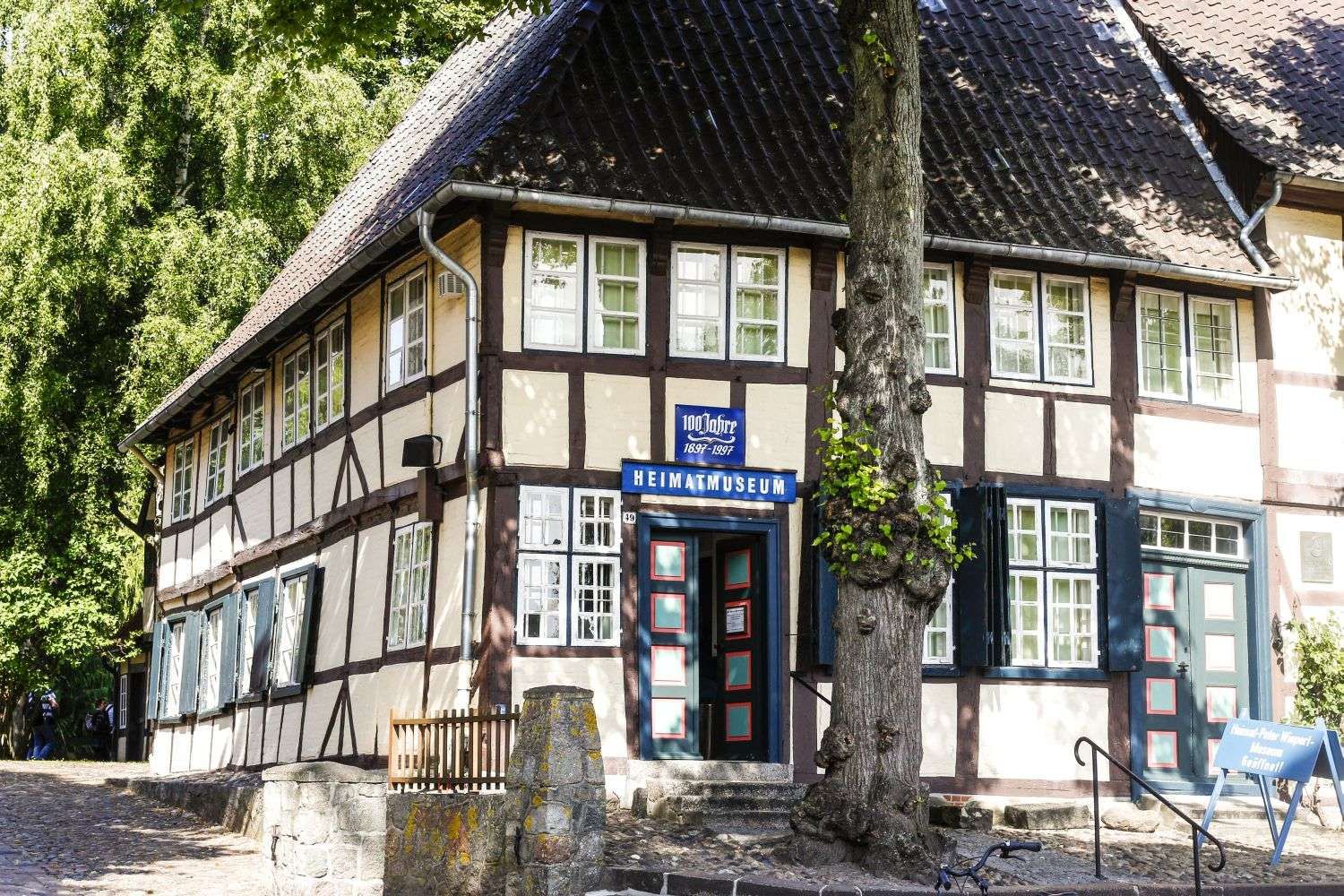 Fachwerkhaus mit Schild 'Heimatmuseum' und Baum im Vordergrund. Das Gebäude hat viele Fenster und eine blaue Tür. Es ist von Bäumen umgeben.