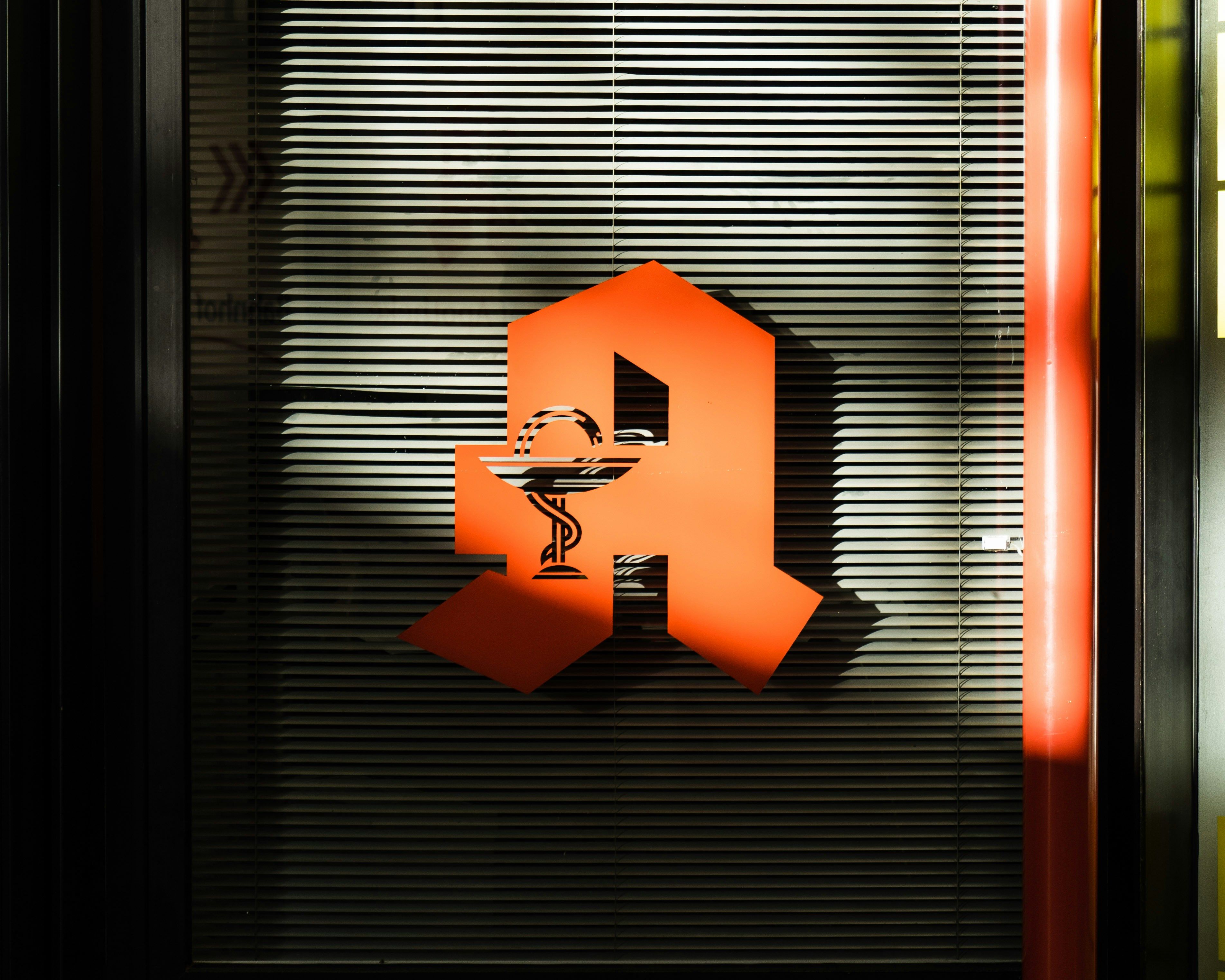 Rotes Apothekenzeichen auf einer Jalousie, beleuchtet von Sonnenlicht. Das Symbol zeigt einen Mörser mit Stößel, typisch für Apotheken.