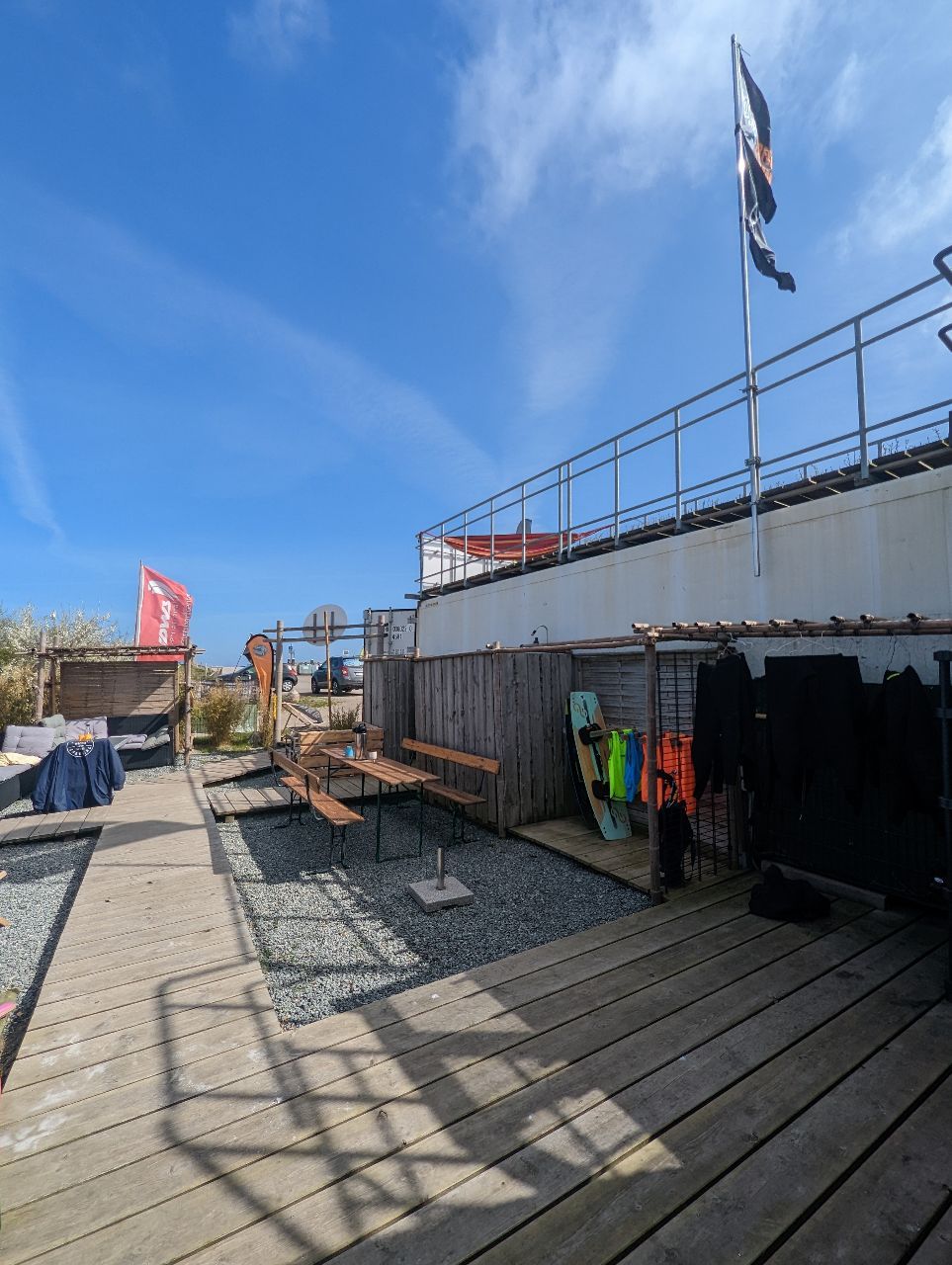 Ein sonniger Außenbereich mit Holzboden, Bänken und Kitesurf-Ausrüstung. Fahnen wehen im Wind, der Himmel ist blau mit leichten Wolken.