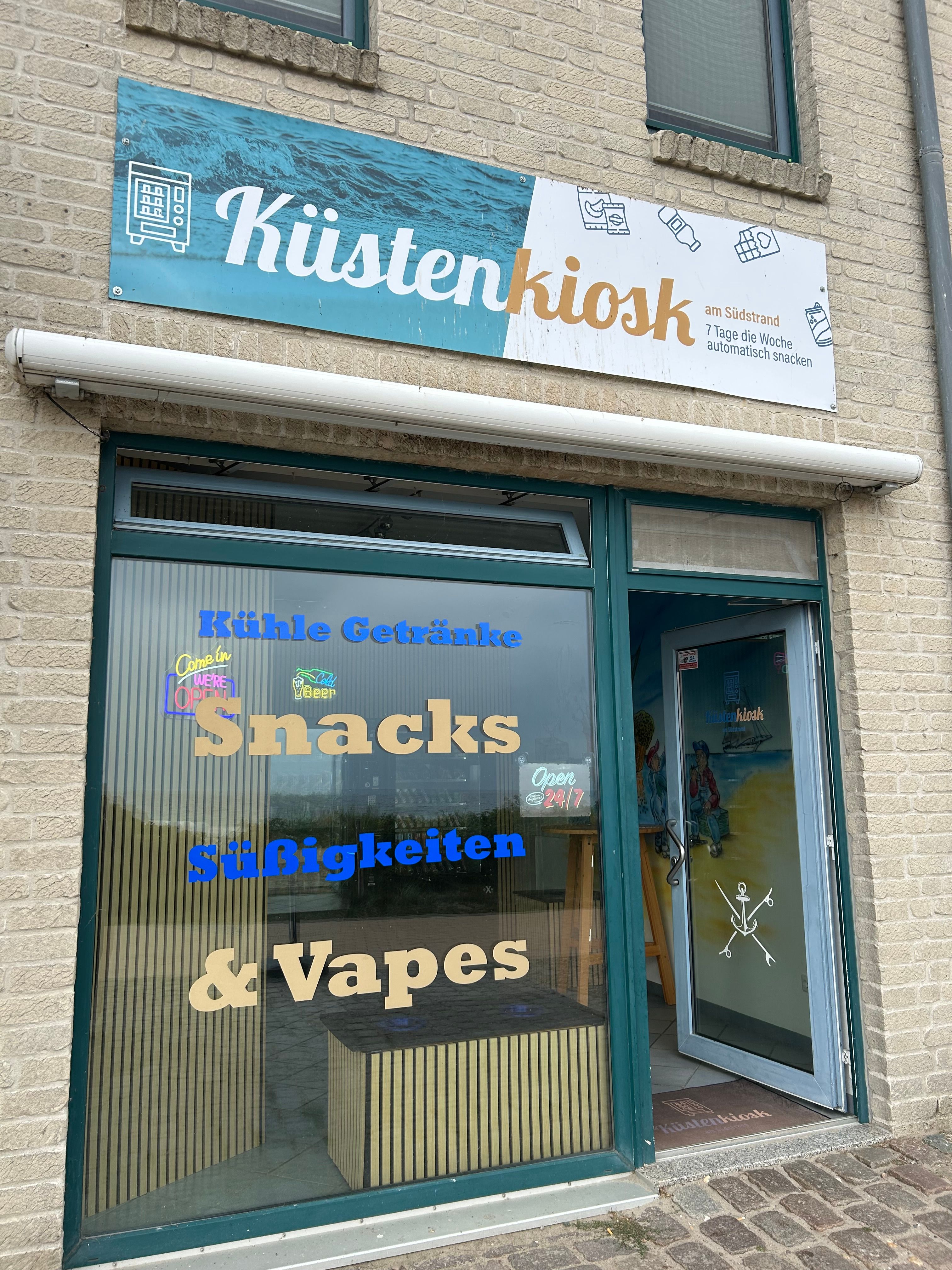 Küstenkiosk mit Schaufenster und Tür. Aufschrift: 'Kühle Getränke, Snacks, Süßigkeiten & Vapes'. Offen 24/7.