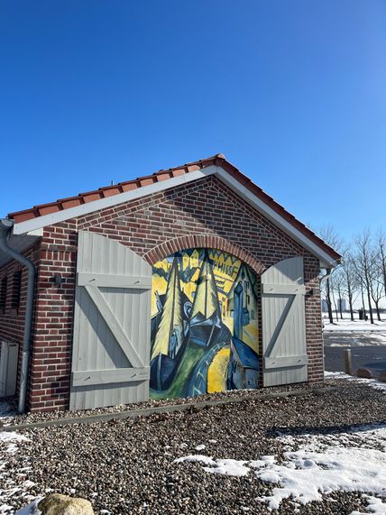 Ein Backsteingebäude mit einem farbenfrohen Wandgemälde von Segelbooten. Der Boden ist mit Kies und Schnee bedeckt, der Himmel ist klar und blau.