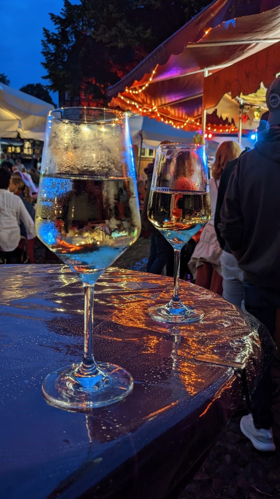 Zwei Weingläser auf einem Tisch bei einem abendlichen Fest. Im Hintergrund sind Lichterketten und Menschen zu sehen.