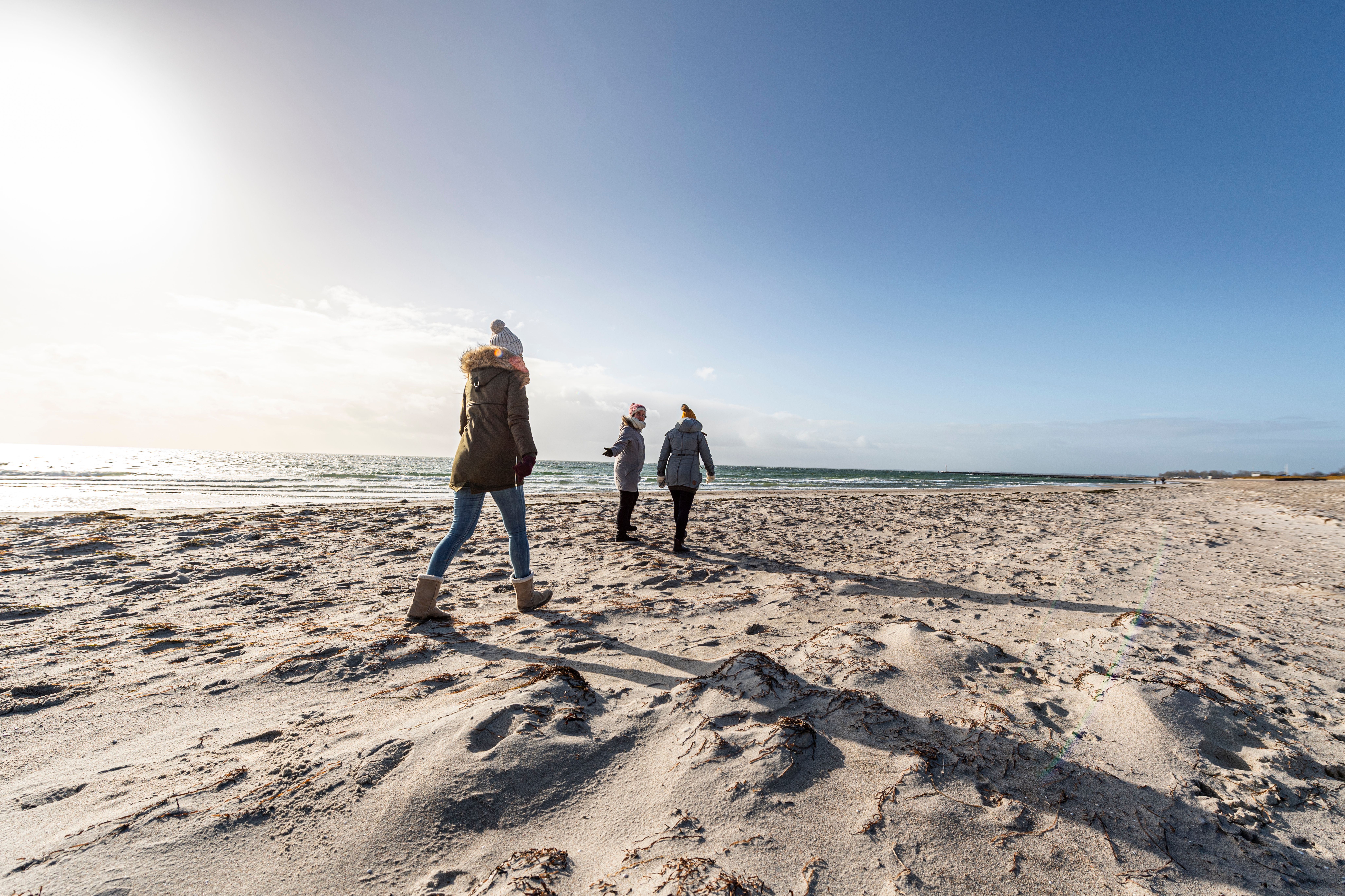 Drei Personen in Winterkleidung gehen am Südstrand auf Fehmarn entlang des sandigen Strands mit Meer im Hintergrund.