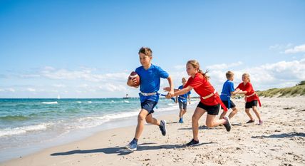 Kinder spielen American Football am Strand. Ein Junge in Blau läuft mit dem Ball, während andere Kinder in Rot versuchen, ihn zu stoppen.