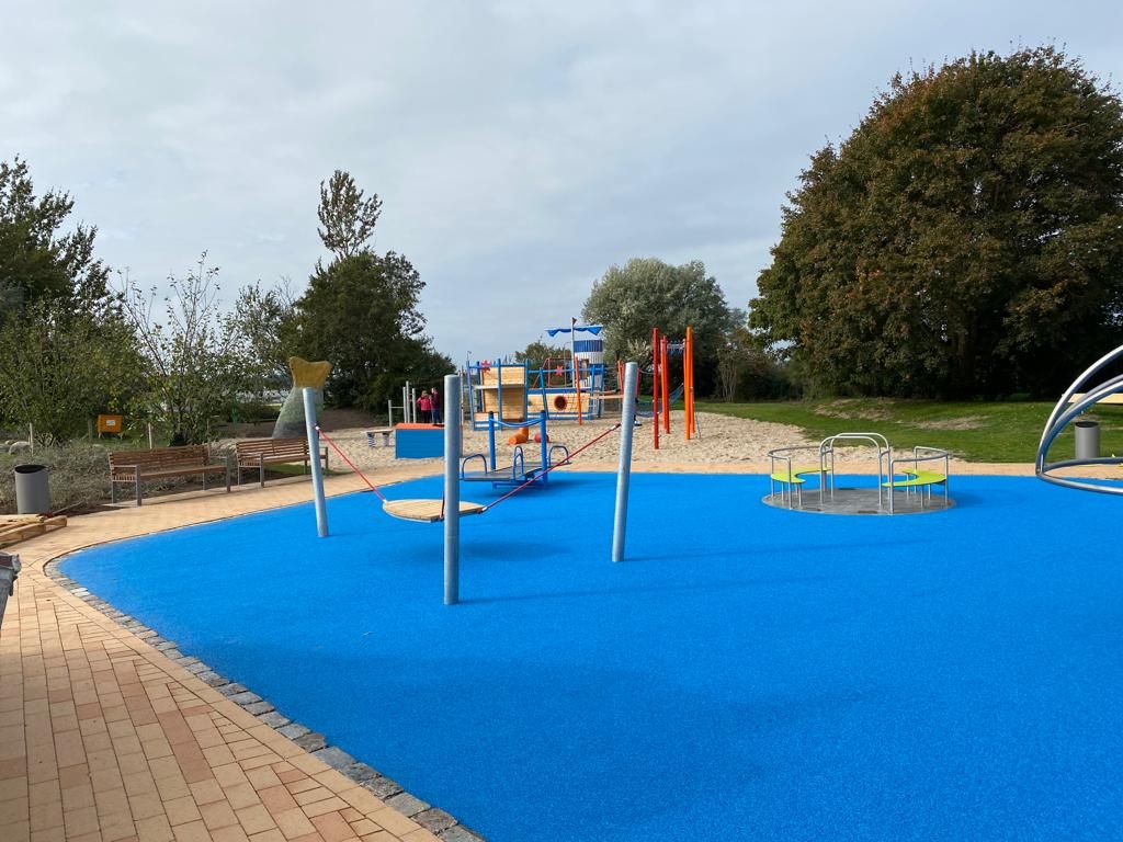 Ein moderner Spielplatz mit blauem Bodenbelag, Klettergerüsten und Bäumen im Hintergrund. Sitzbänke und ein Weg sind ebenfalls zu sehen.