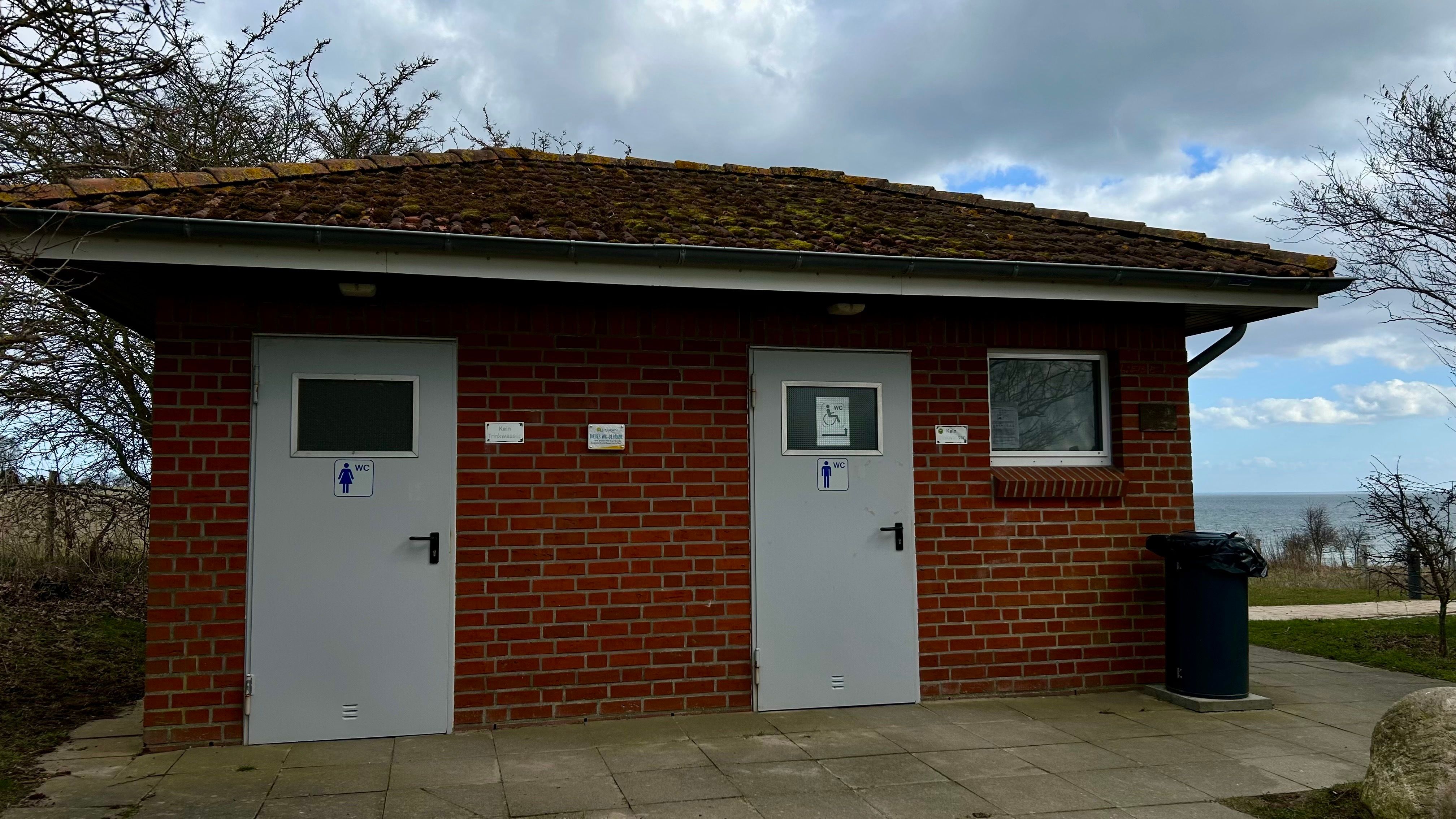 Backsteingebäude mit zwei Toilettentüren, umgeben von Bäumen und mit Blick auf das Meer. Der Himmel ist bewölkt.