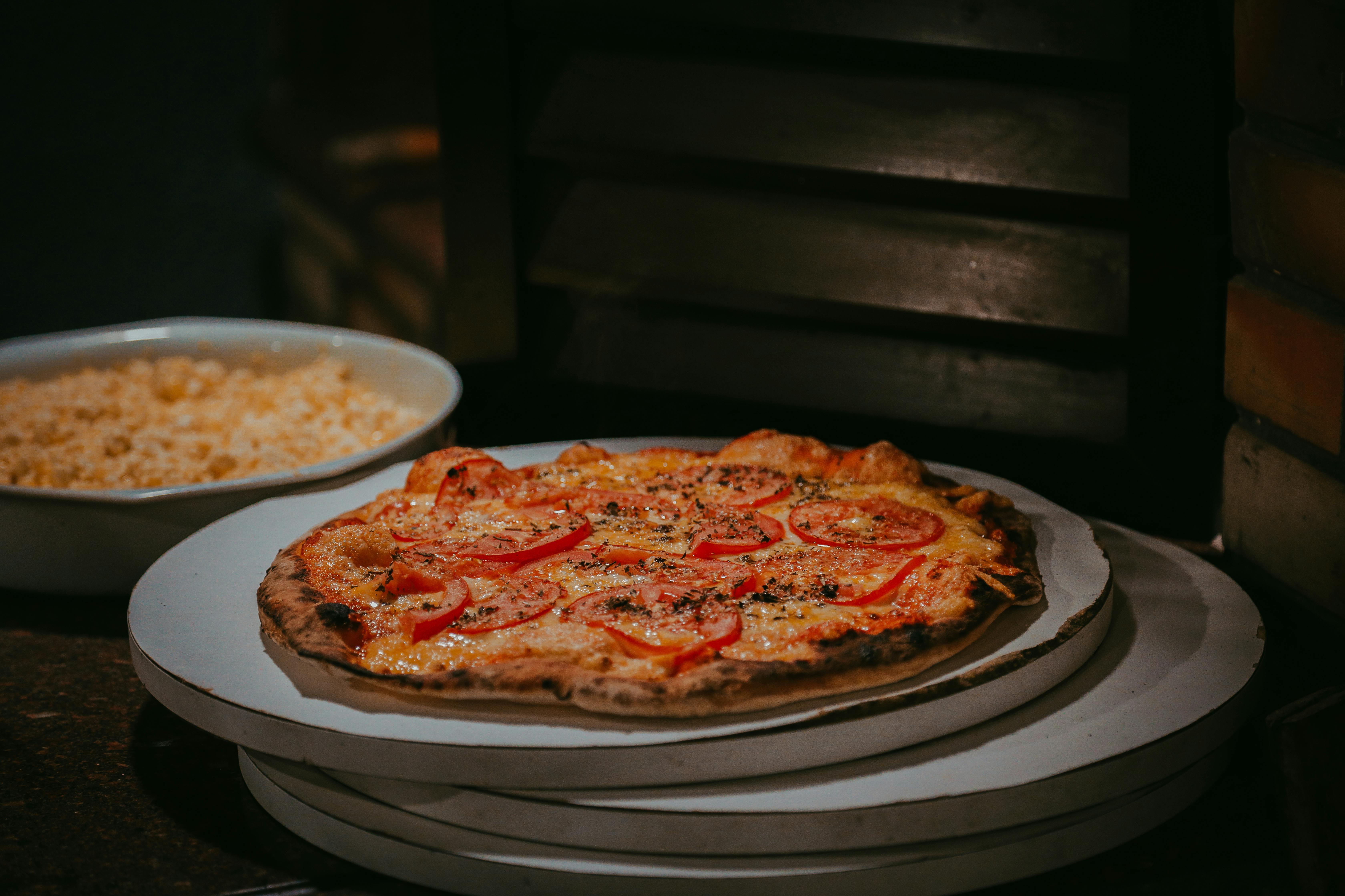 Eine Pizza mit Tomaten und Käse auf einem Teller, daneben eine Schüssel mit Mais. Die Szene ist in warmem Licht gehalten.