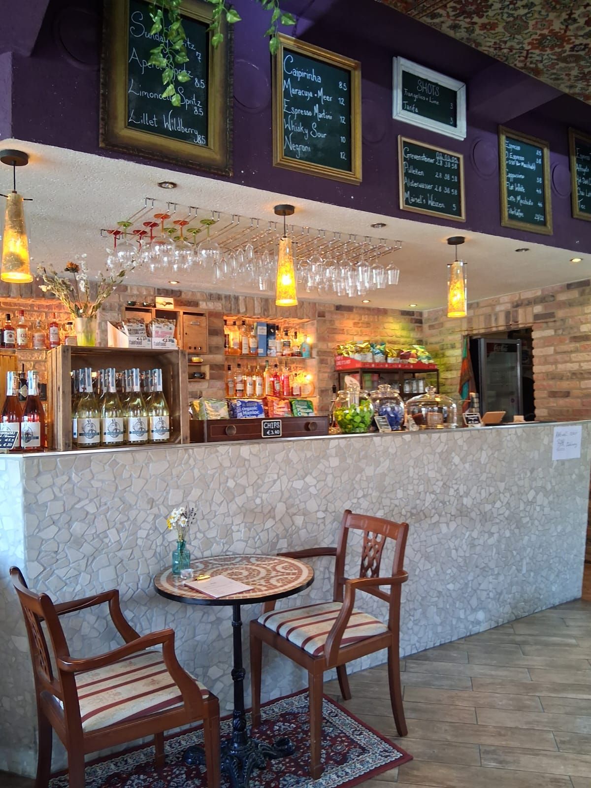 Gemütliches Café mit Bar, dekorativen Tafeln und hängenden Gläsern. Zwei Stühle und ein Tisch stehen auf einem Teppich.