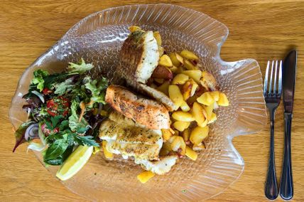 Ein Fischgericht auf einem gläsernen Teller in Fischform. Es enthält gebratenen Fisch, Bratkartoffeln und einen frischen Salat mit Zitrone.