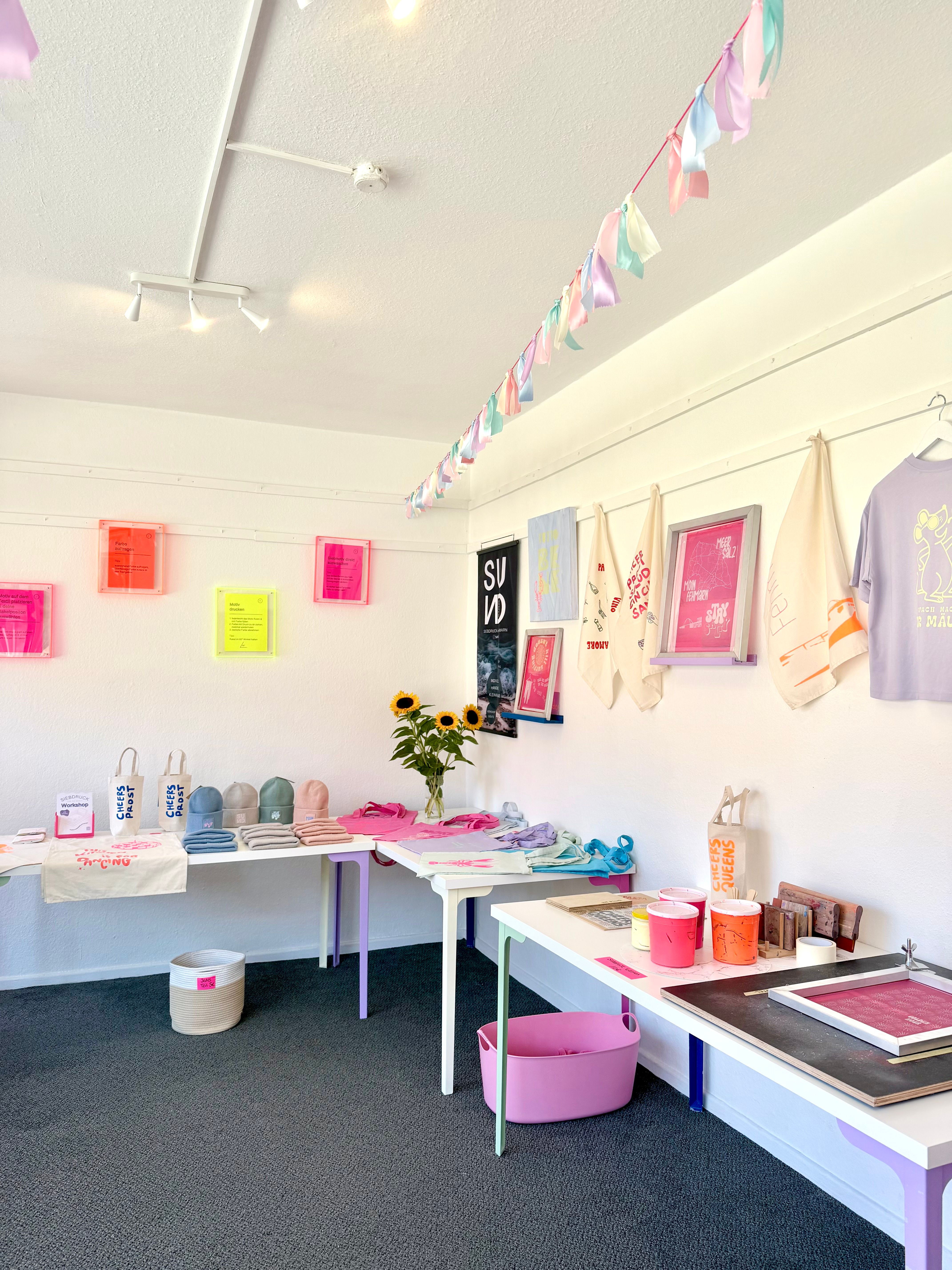 Ein heller Raum mit bunten Dekorationen und kreativen Produkten auf Tischen. An den Wänden hängen Kunstwerke und Stofftaschen. Eine Girlande schmückt die Decke.
