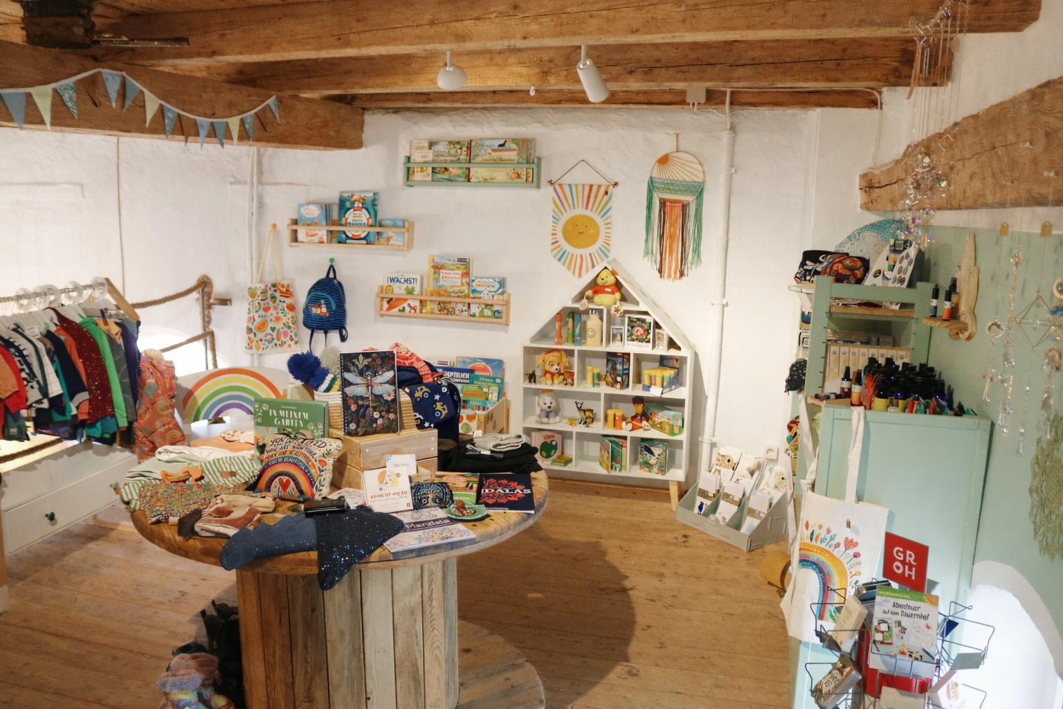 Ein gemütlicher Hofladen mit bunten Kinderkleidern, Spielzeug und Dekorationen. Holzbalken und warme Farben schaffen eine einladende Atmosphäre.