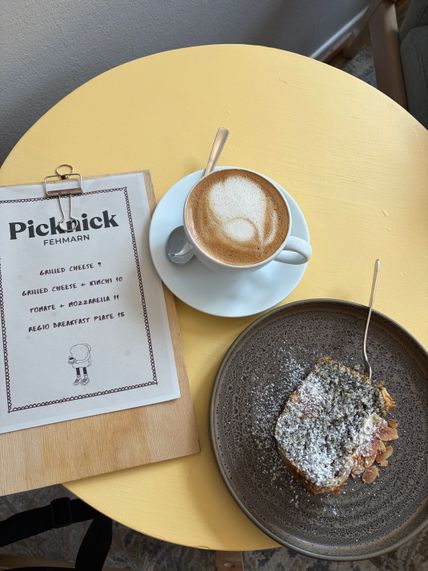 Auf einem gelben Tisch stehen eine Tasse Cappuccino, ein Stück Kuchen auf einem Teller und eine Speisekarte von Picknick Fehmarn.