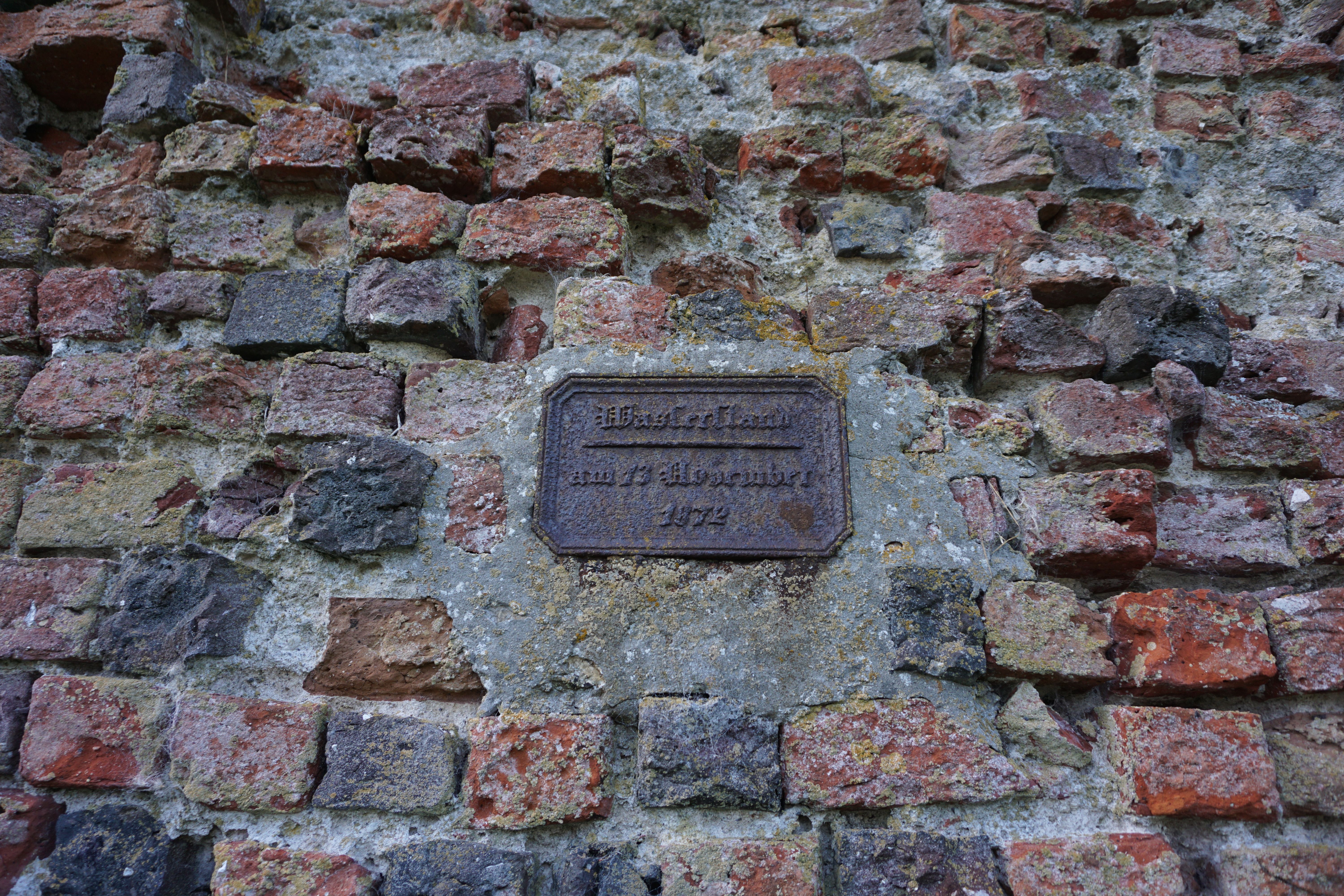 Eisenplatte an einer Mauer mit der Inschrift 'Wasserstand, 13. November 1872', umgeben von roten und grauen Ziegelsteinen.