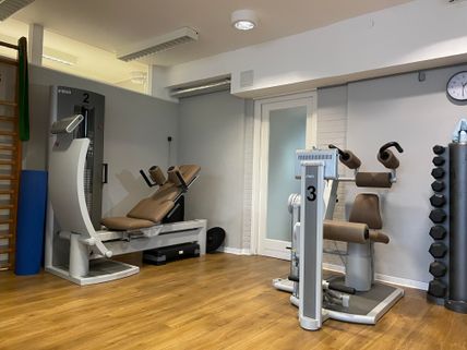 Ein Fitnessstudio mit modernen Trainingsgeräten und Hanteln auf einem Holzfußboden. Die Geräte sind nummeriert und es gibt eine Uhr an der Wand.