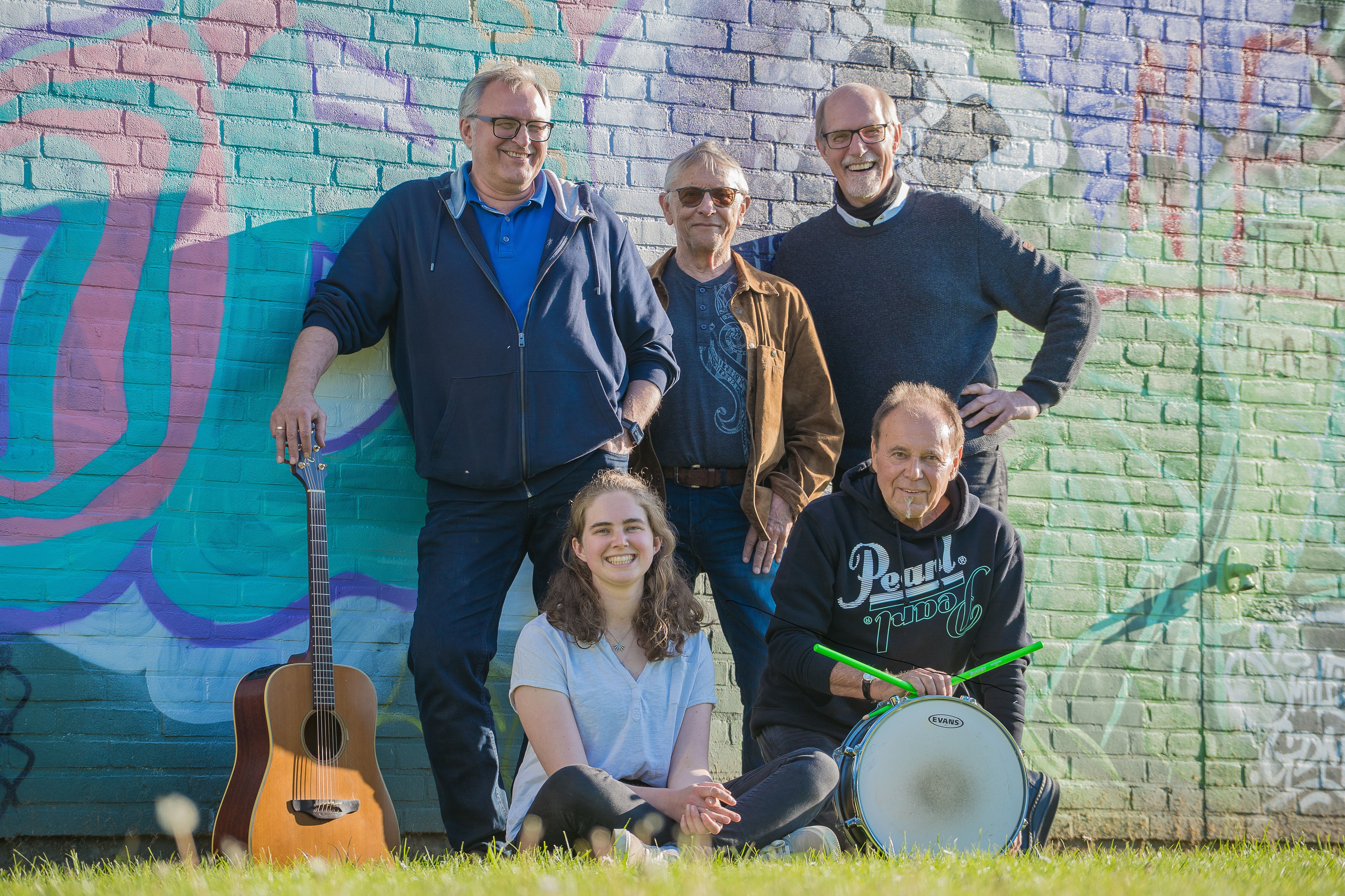 Fünf Personen vor einer bunten Graffiti-Wand. Eine Frau sitzt vorne, vier Männer stehen oder hocken dahinter. Eine Gitarre und eine Trommel sind sichtbar.