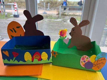 Zwei selbstgebastelte Osterkörbe aus Papier, dekoriert mit Hasen, Eiern und Blumen. Einer trägt die Aufschrift 'Frohe Ostern'.