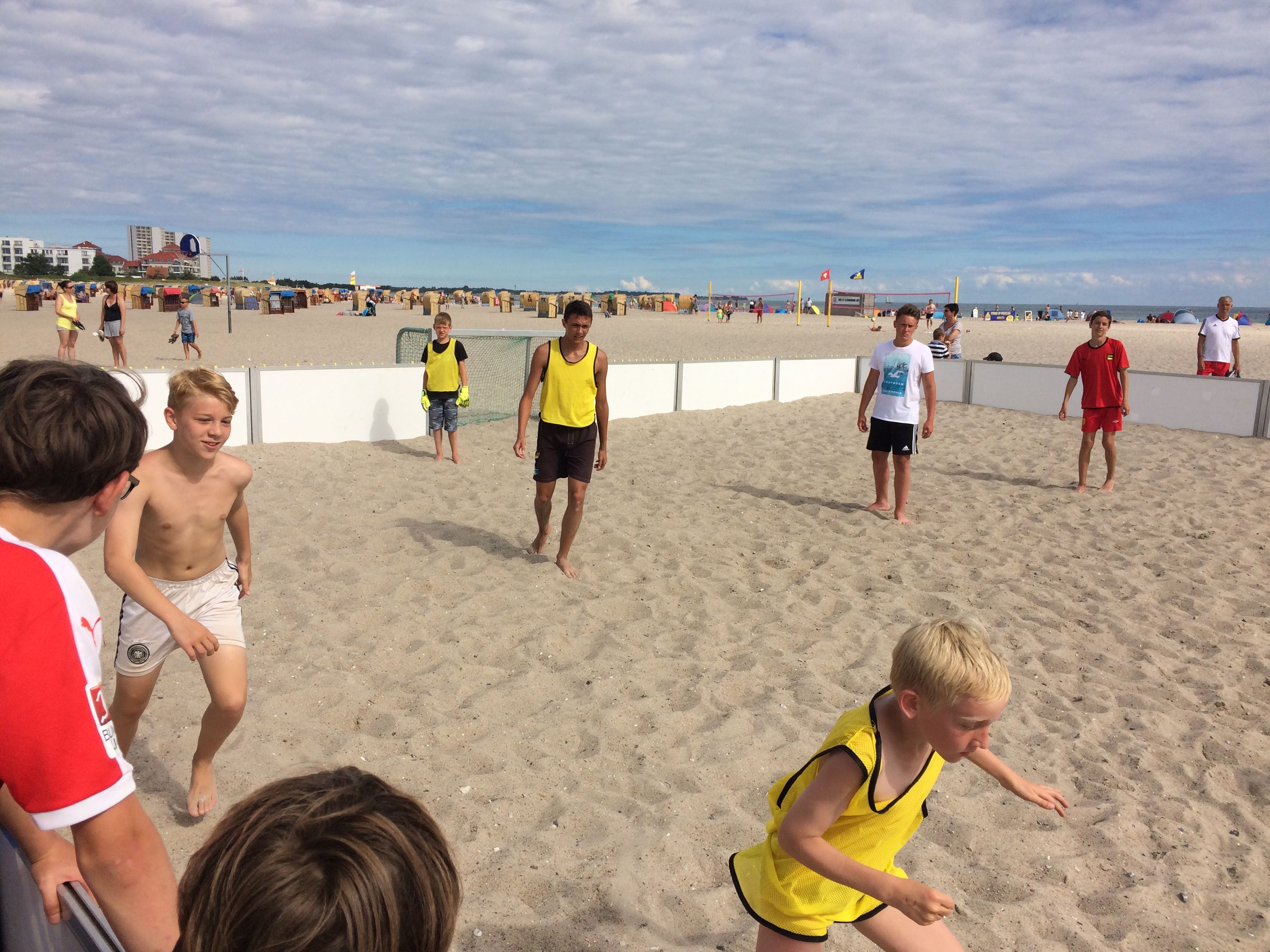 Kinder spielen Beachsoccer am Strand. Einige tragen gelbe Leibchen. Im Hintergrund sind Strandkörbe und ein bewölkter Himmel zu sehen.