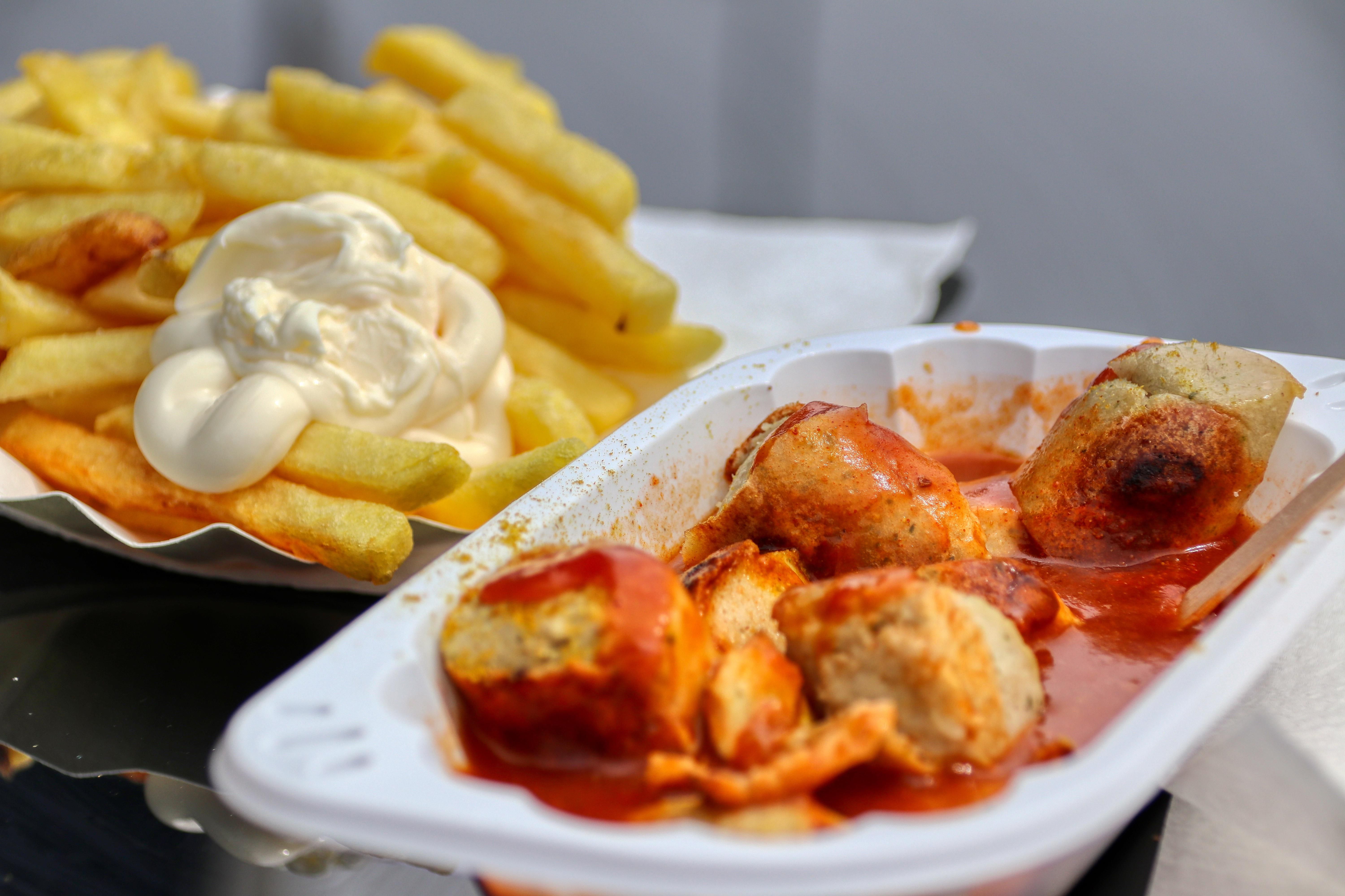 Currywurst mit würziger Soße in einer Schale, daneben Pommes mit Mayonnaise. Ein klassisches deutsches Fast-Food-Gericht.