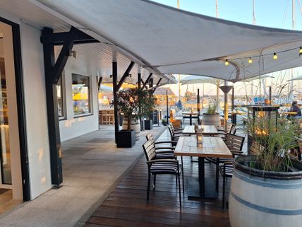 Gemütlicher Außenbereich eines Restaurants am Hafen von Lemkenhafen mit Holztischen, Stühlen und Segeltuchüberdachung. Im Hintergrund sind Segelboote zu sehen.