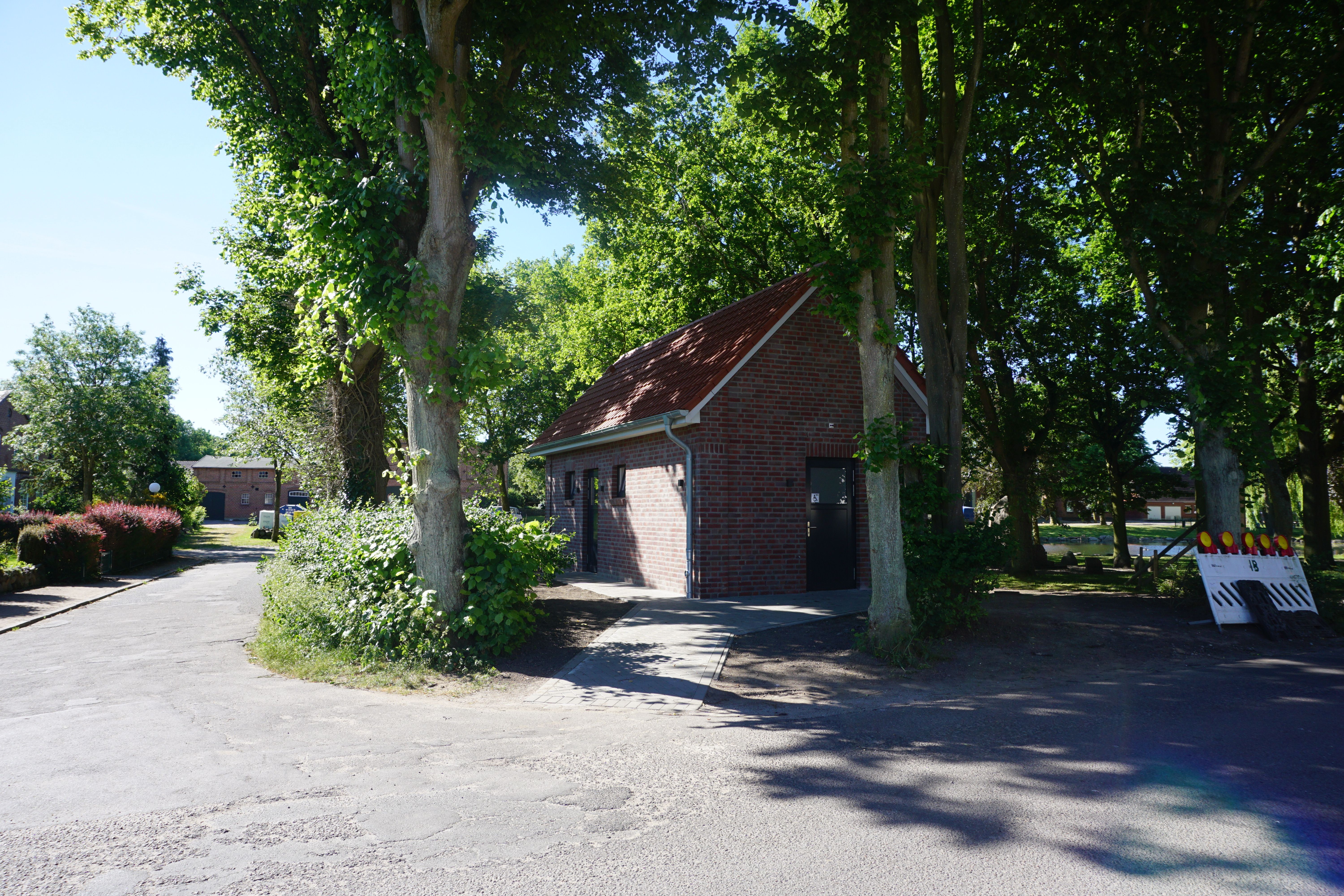 Ein kleines Backsteingebäude mit rotem Dach steht zwischen hohen Bäumen. Ein asphaltierter Weg führt daran vorbei. Im Hintergrund sind weitere Gebäude zu sehen.