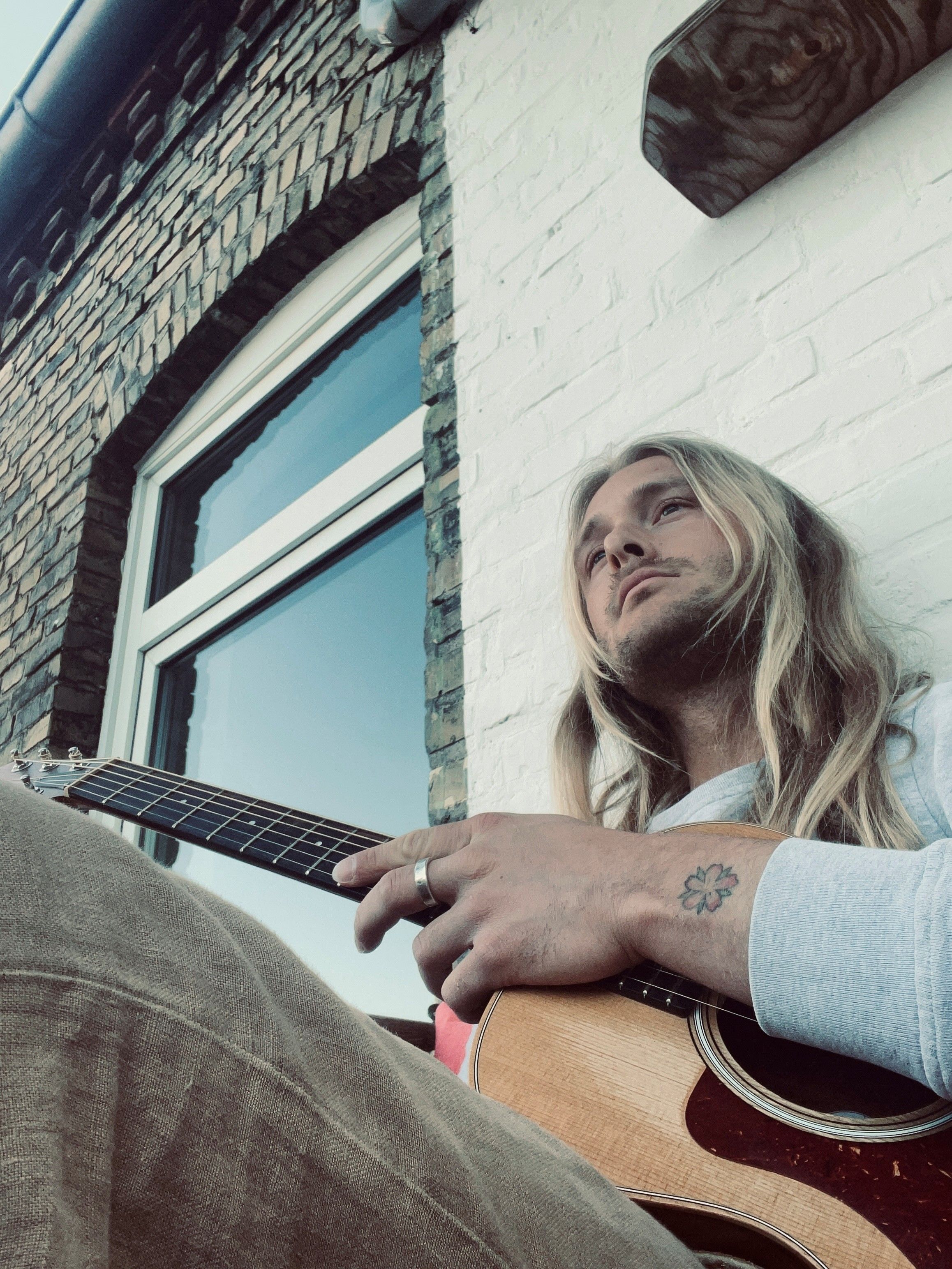 Eine Person mit langen Haaren sitzt entspannt vor einem Backsteingebäude und spielt Gitarre. Ein Tattoo ist auf der Hand sichtbar.