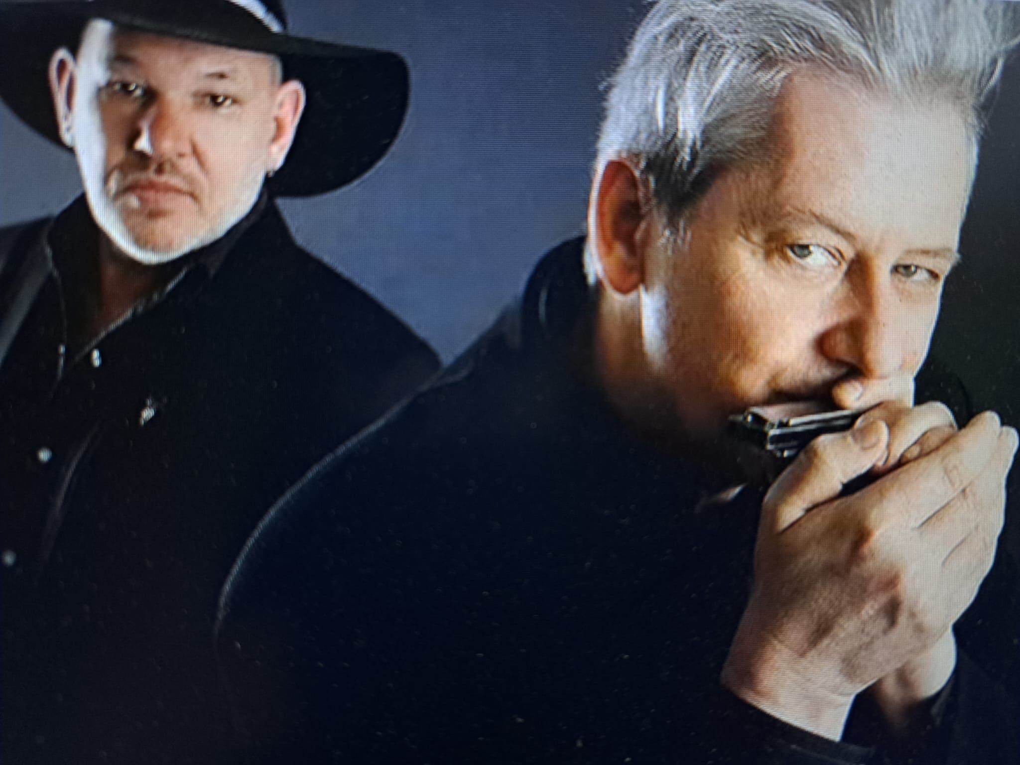 Zwei Männer in dunkler Kleidung, einer spielt Mundharmonika, der andere trägt einen Hut und schaut in die Kamera.