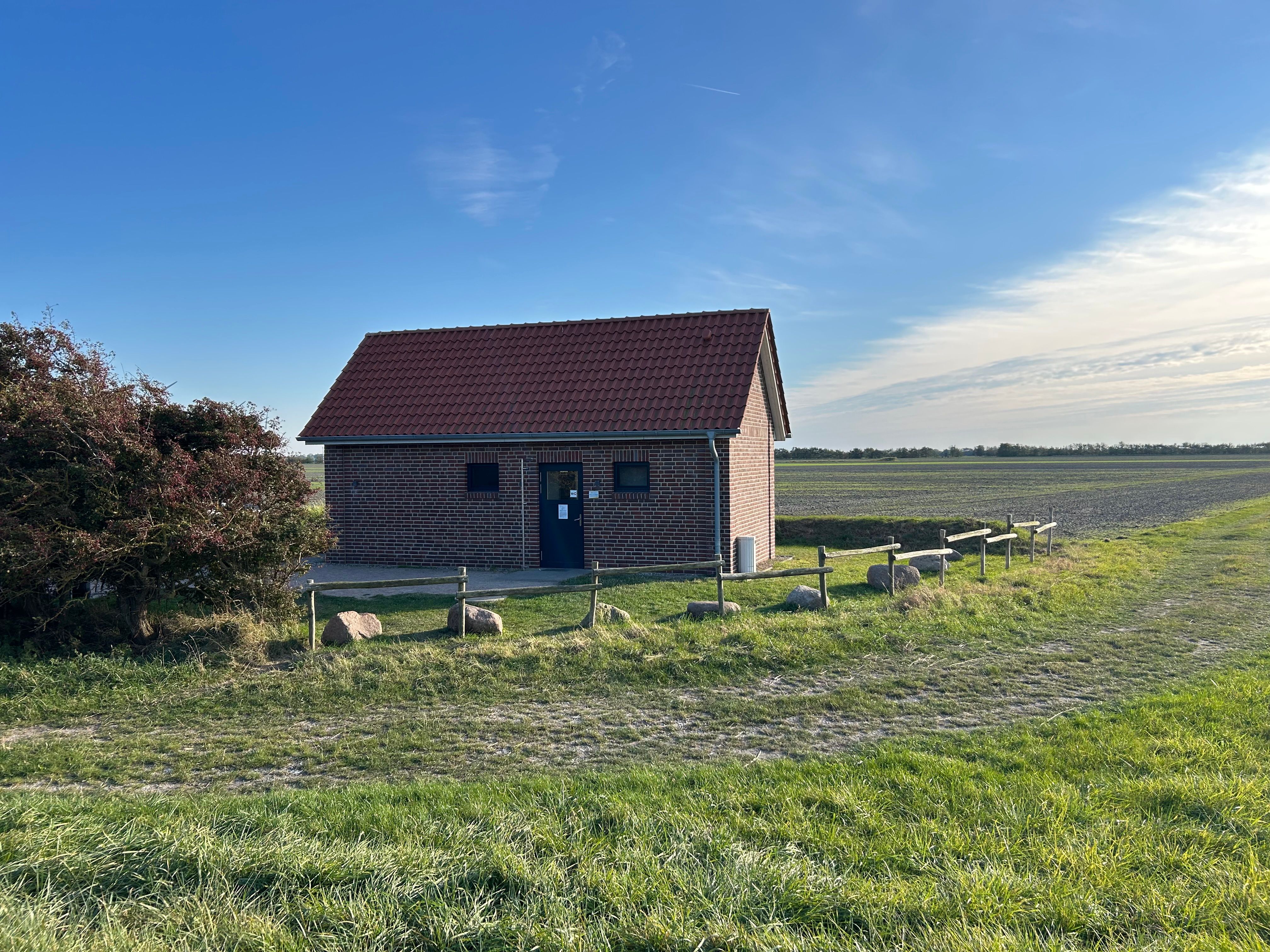 Ein kleines Backsteingebäude mit rotem Ziegeldach steht auf einem Feld unter blauem Himmel. Ein Weg führt daran vorbei, umgeben von Gras und Büschen.
