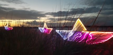 Leuchtende Papierboote in verschiedenen Farben stehen in einer Wiese bei Sonnenuntergang. Der Himmel ist bewölkt und die Boote strahlen hell.