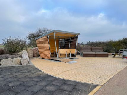 Moderne Wetterschutzhütte aus Holz mit Sitzgelegenheiten auf einem gepflasterten Platz, umgeben von Büschen und Steinen.