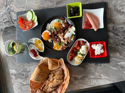 Vielfältiges Frühstück mit Spiegelei, Wurst, Brot, Tomaten, Gurken, Oliven, Aufschnitt, Marmelade und einem Getränk mit Zitrone und Minze.