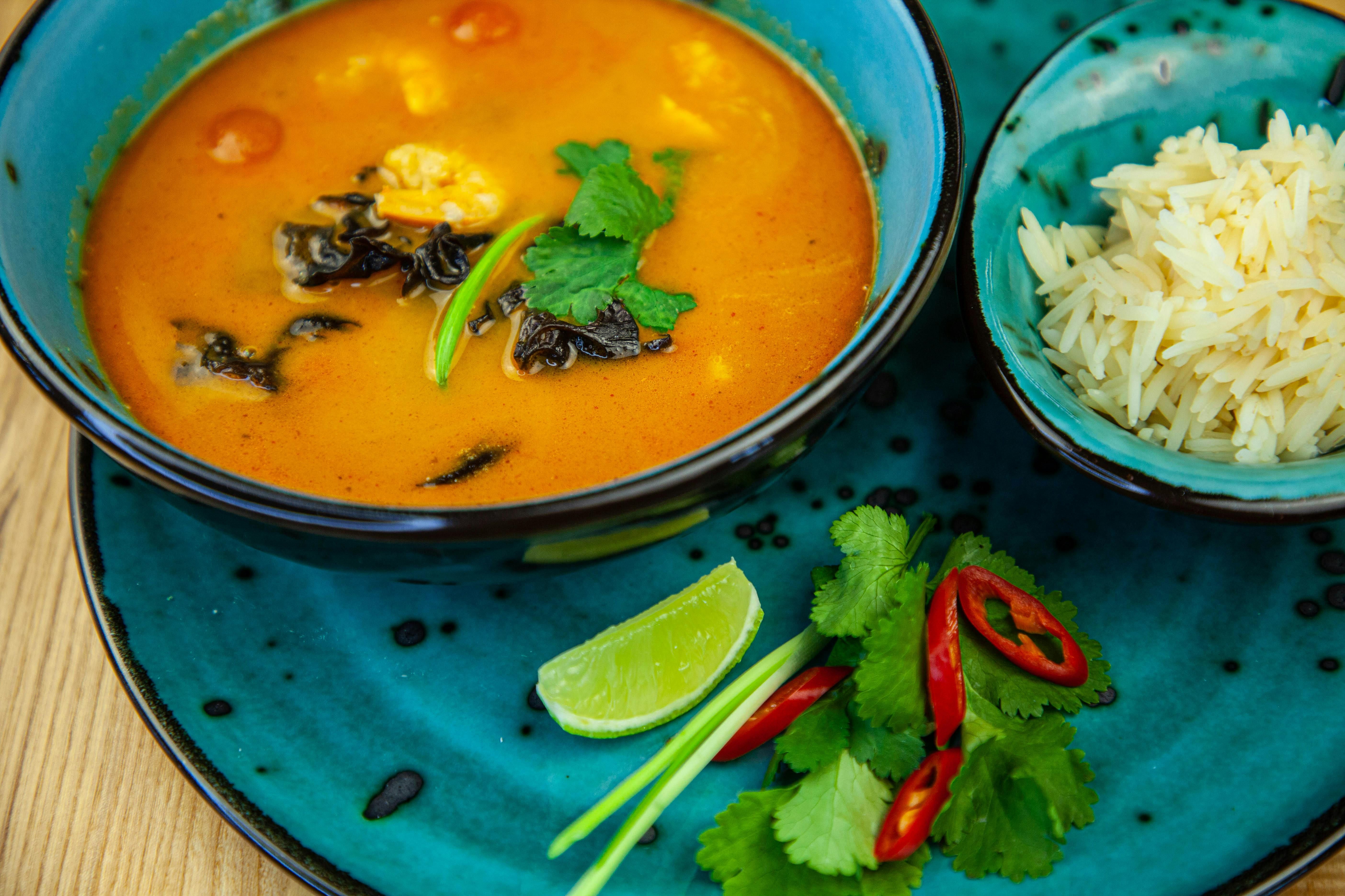 Rote Suppe mit Koriander und Pilzen in einer Schüssel, daneben Reis und Beilagen wie Limette und Chili auf einem blauen Teller.