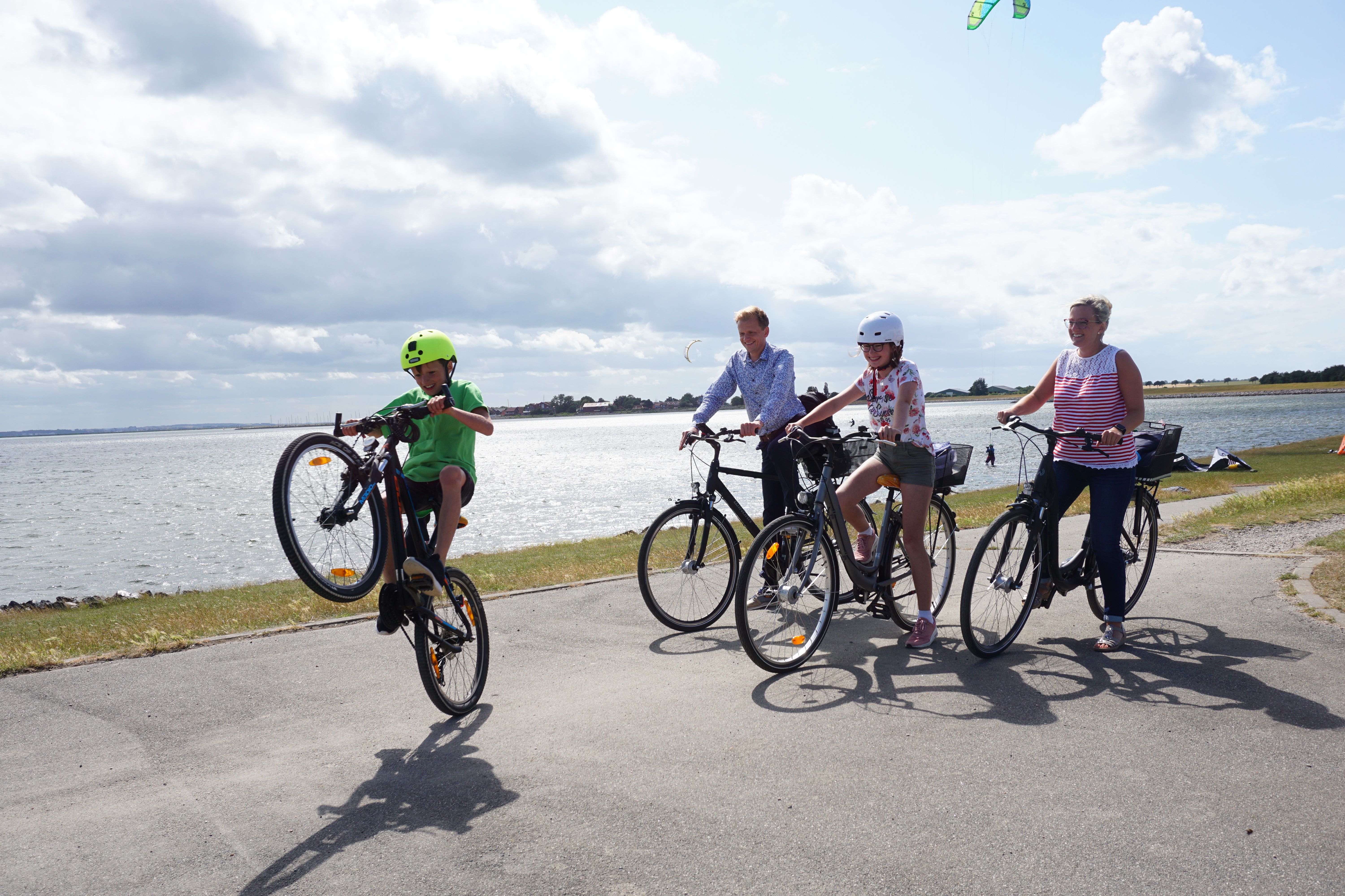 Eine Familie radelt fröhlich auf einem Deich entlang, während ein Kind einen Wheelie macht. Im Hintergrund ist das Meer zu sehen.