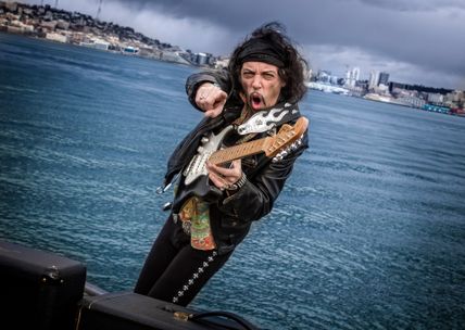 Ein Gitarrist in Lederjacke spielt leidenschaftlich E-Gitarre vor einer Küstenkulisse. Der Himmel ist bewölkt, und im Hintergrund sind Gebäude zu sehen.