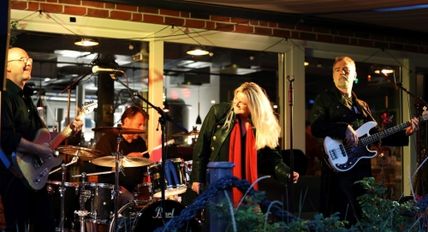 Eine Band performt auf einer Bühne. Die Sängerin trägt einen roten Schal, begleitet von Gitarrist, Schlagzeuger und Bassist.