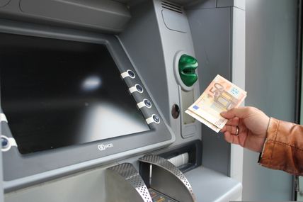 Eine Hand hält zwei 50-Euro-Scheine vor einem Geldautomaten mit einem schwarzen Bildschirm.