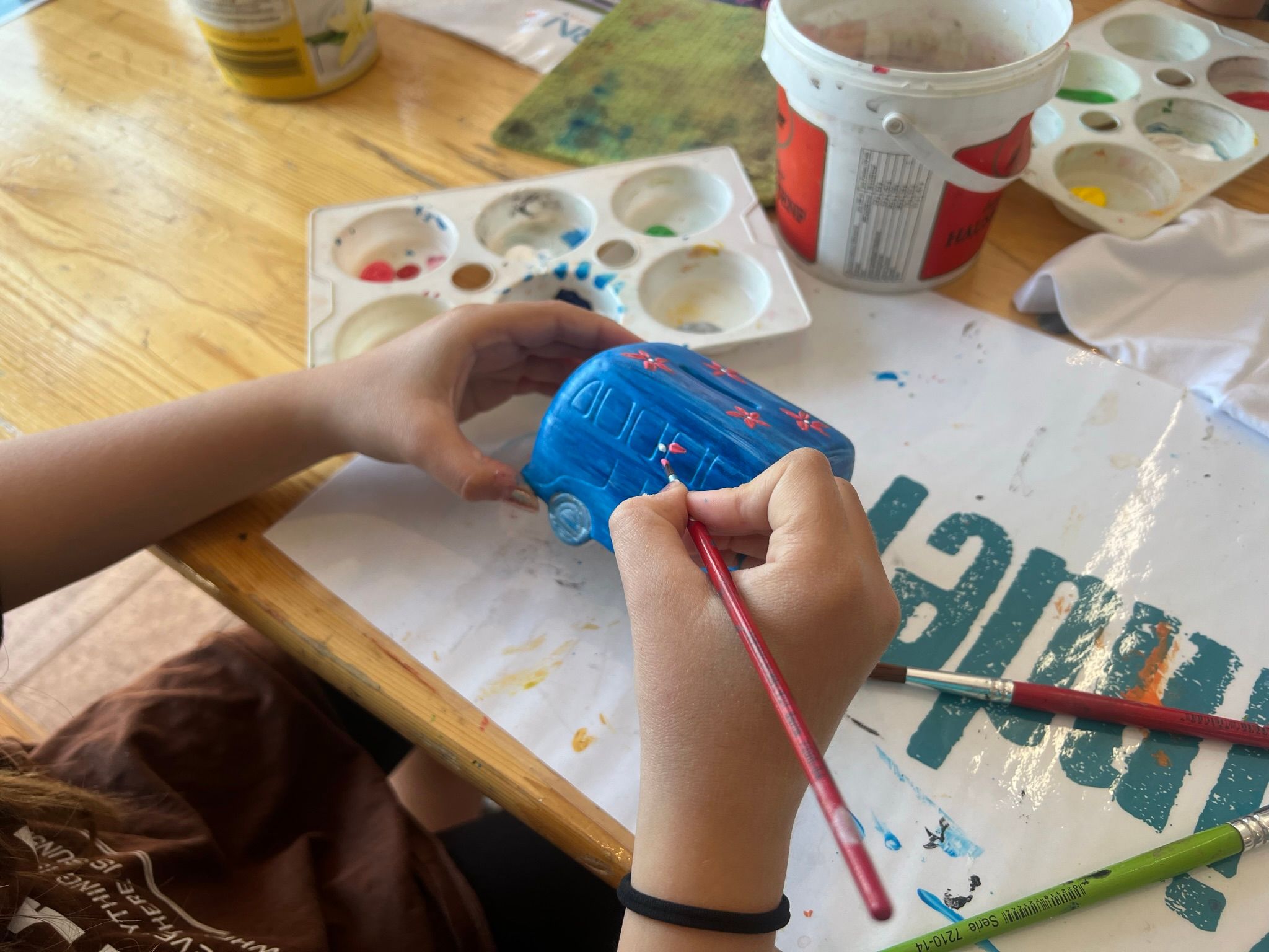Eine Person bemalt ein blaues Porzellanmodell eines Autos mit Blumenmustern. Auf dem Tisch liegen Farben und Pinsel.