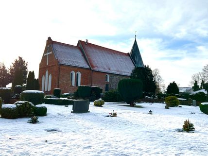 Eine Kirche mit rotem Ziegeldach und grünem Turm steht auf einem verschneiten Friedhof. Die Gräber sind mit Schnee bedeckt, und der Himmel ist leicht bewölkt.