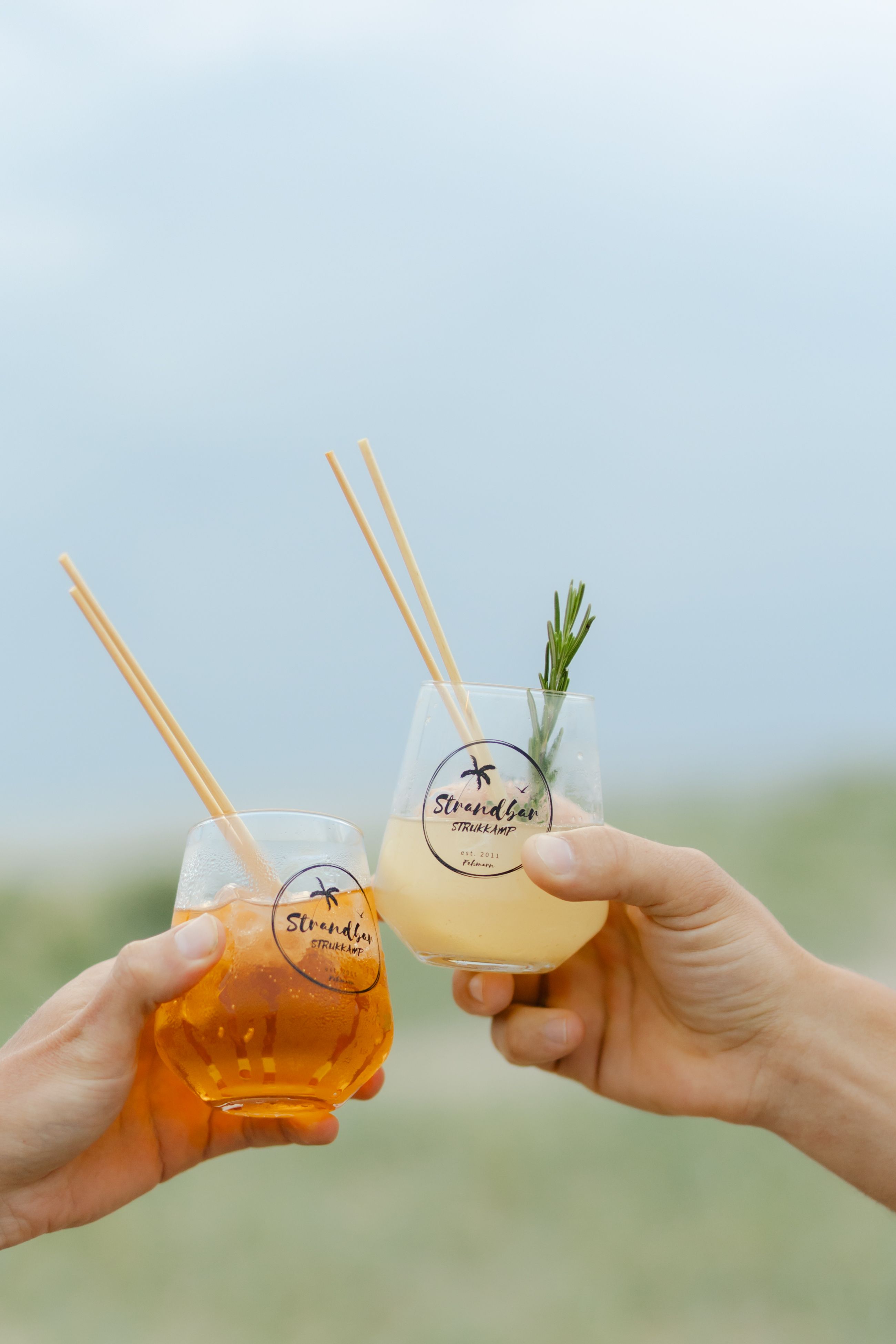 Zwei Personen stoßen mit Cocktails in Gläsern der Strandbar Strukkamp an. Im Hintergrund ist ein bewölkter Himmel zu sehen.