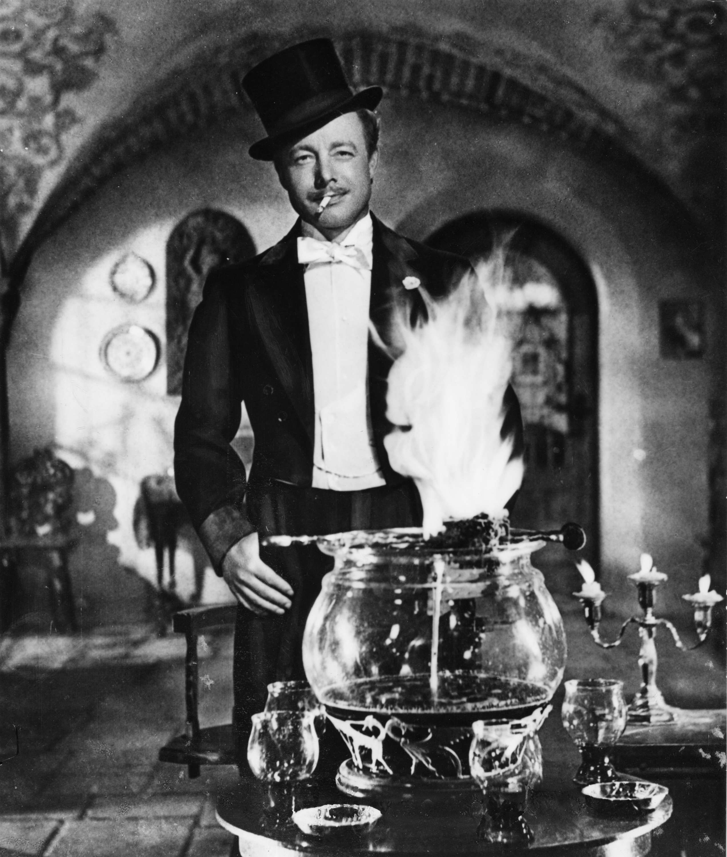 Ein Mann im Anzug mit Zylinder steht hinter einer brennenden Feuerzangenbowle in einem stilvollen Raum.