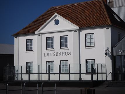 Das Lotsenhus in Burgstaaken ist ein weißes Gebäude mit rotem Ziegeldach und der Aufschrift 'Lotsenhus'. Es steht unter klarem, blauem Himmel.