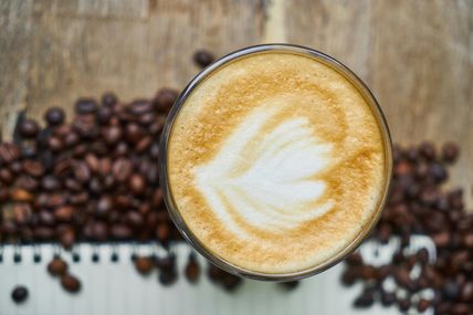 Ein Cappuccino mit Latte Art in Form eines Blattes, umgeben von Kaffeebohnen auf einem Holztisch. Perfekt für Kaffeeliebhaber.