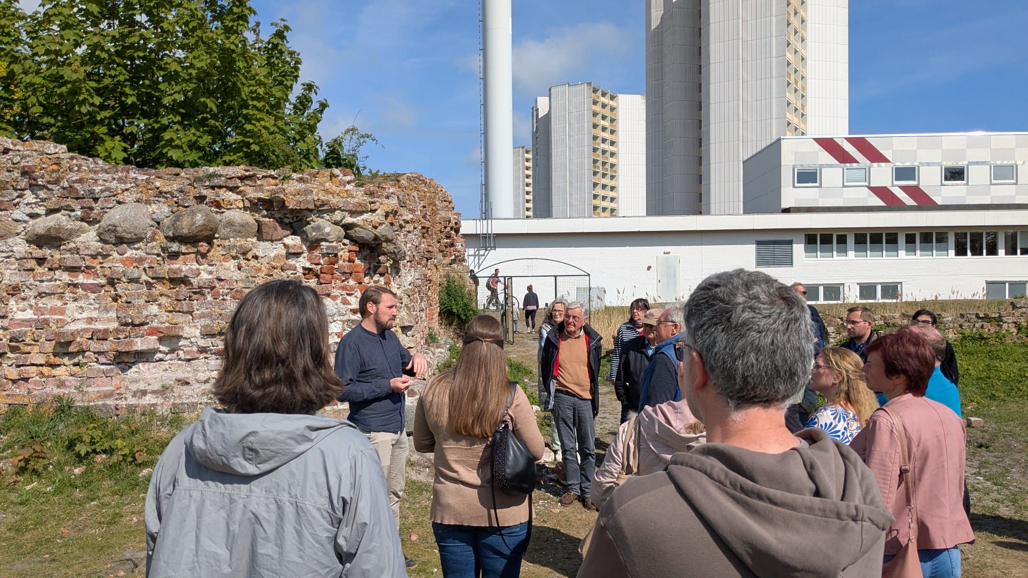 Eine Gruppe von Menschen bei einer Führung vor einer alten Mauer. Im Hintergrund sind moderne Gebäude und ein blauer Himmel zu sehen.