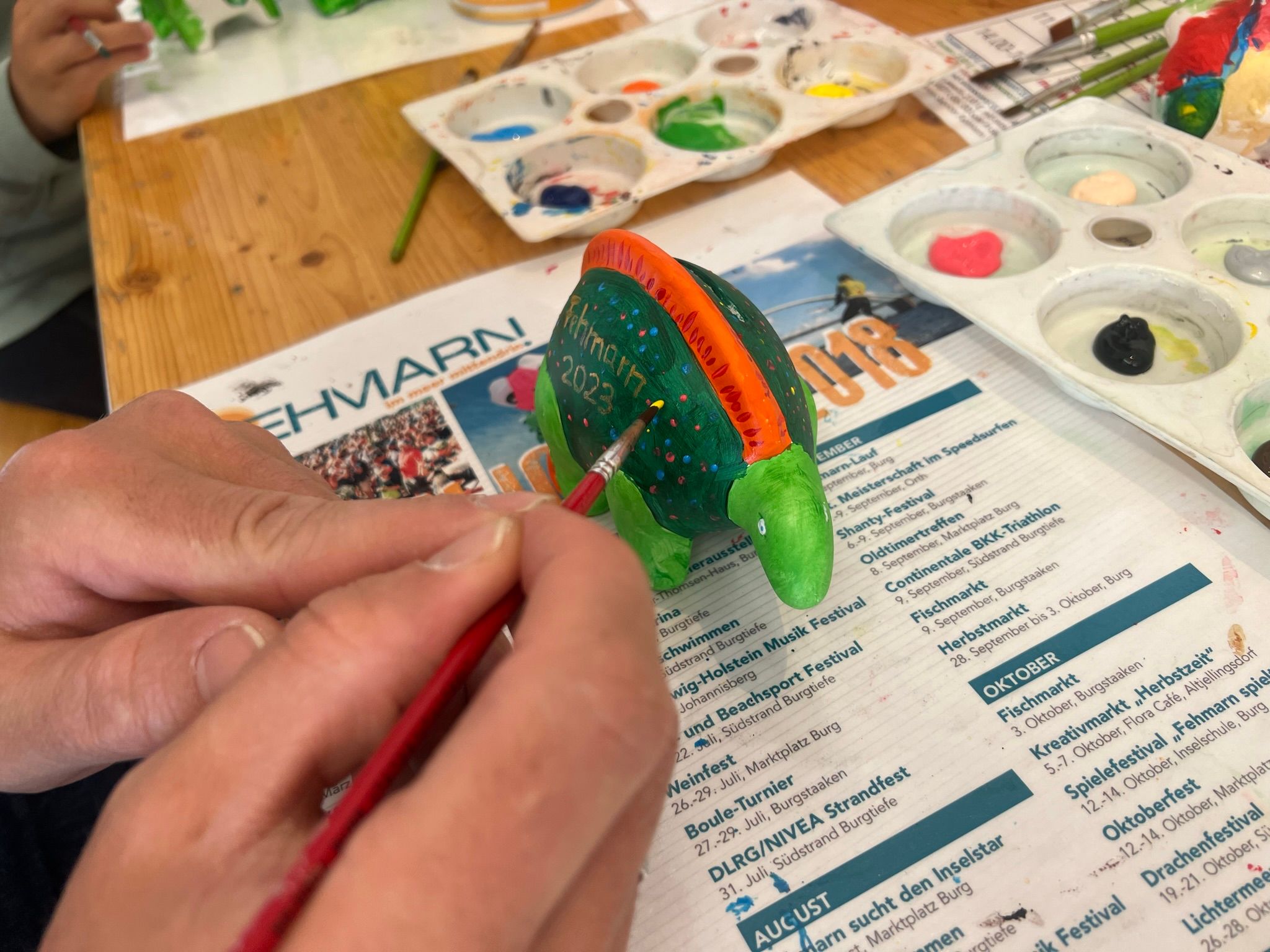 Eine Person bemalt eine grüne Porzellanfigur mit einem Pinsel. Im Hintergrund sind Farbpaletten und ein Kalenderblatt zu sehen.