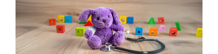 Ein lila Plüschhase sitzt auf einem Holzboden, umgeben von bunten Holzklötzen. Vor ihm liegt ein Stethoskop.