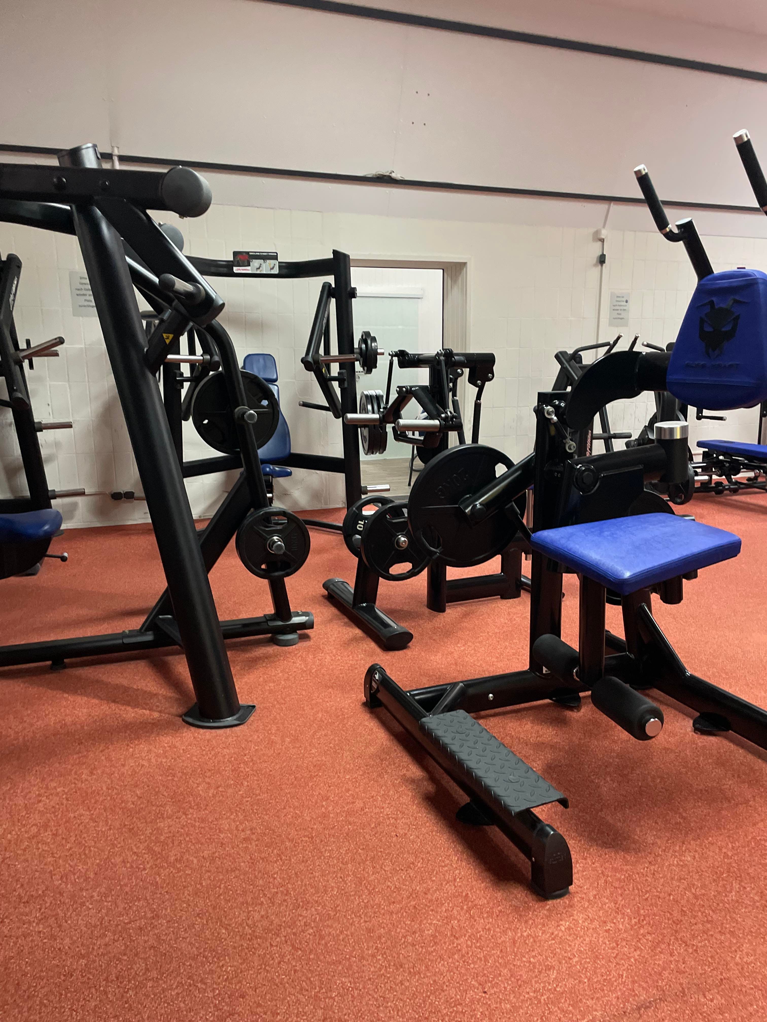 Ein Fitnessstudio mit verschiedenen Krafttrainingsgeräten auf rotem Boden. Die Geräte sind schwarz mit blauen Polstern.