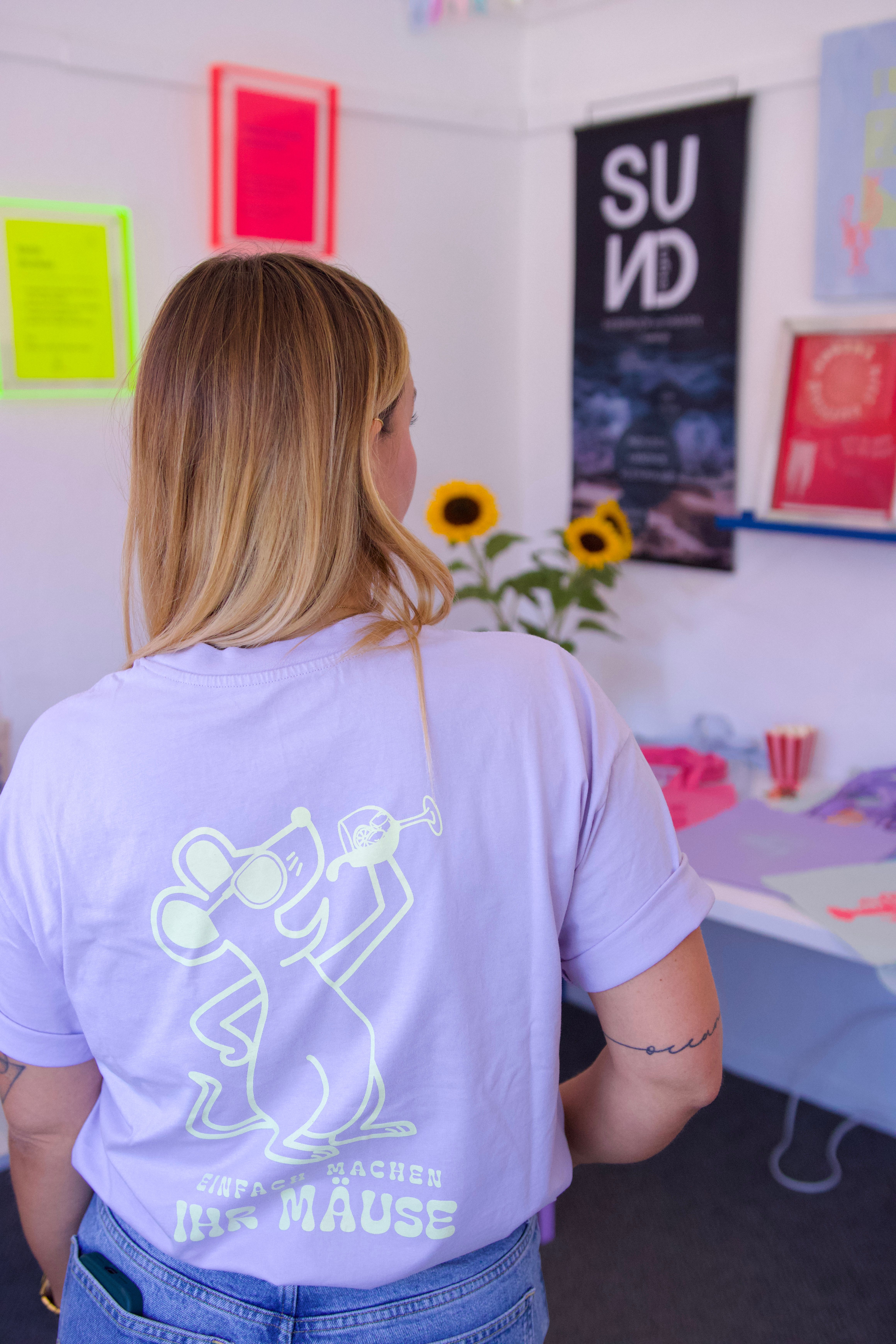 Eine Frau trägt ein T-Shirt mit einer Mausgrafik und dem Text 'Einfach machen ihr Mäuse'. Im Hintergrund sind bunte Poster und Sonnenblumen zu sehen.