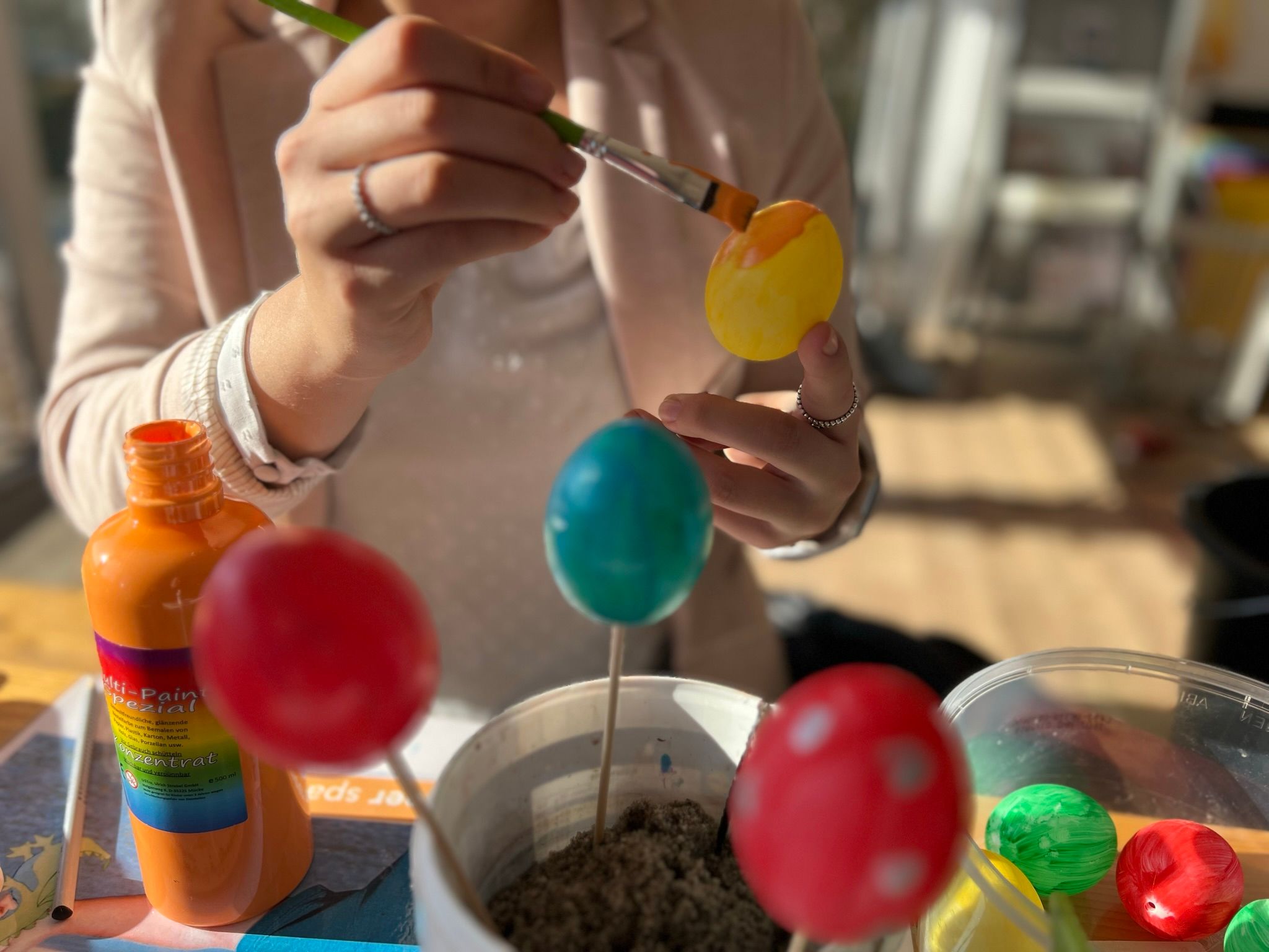 Eine Person bemalt ein gelbes Osterei mit orangefarbener Farbe. Im Vordergrund stehen weitere bunte Eier auf Stäben.