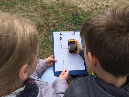Zwei Kinder beim Geocaching: Sie verwenden ein GPS-Gerät und notieren Koordinaten auf einem Klemmbrett.