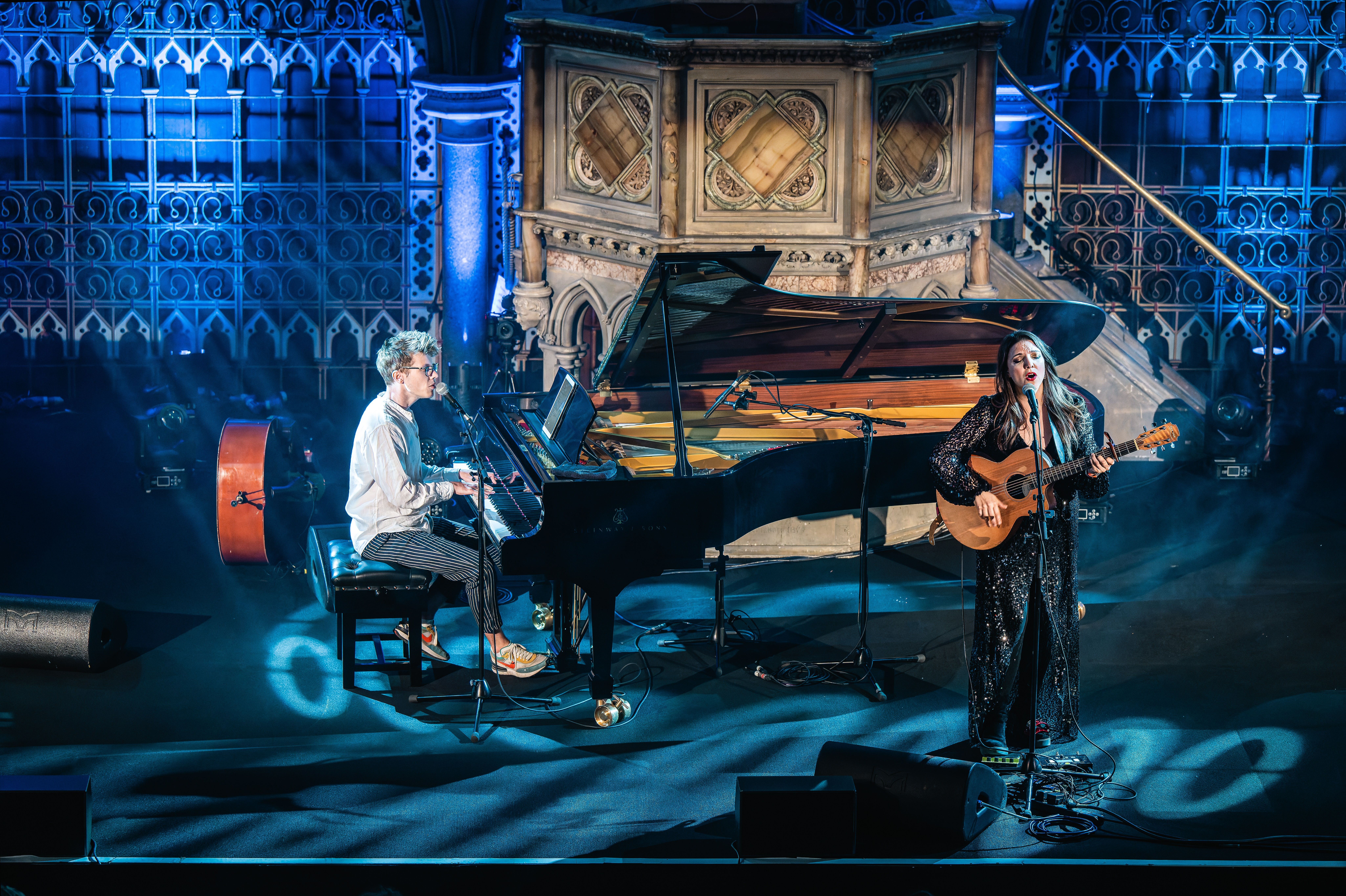 Zwei Musiker auf einer Bühne, einer am Klavier, der andere mit Gitarre. Die Bühne ist stimmungsvoll beleuchtet, im Hintergrund sind dekorative Wände zu sehen.