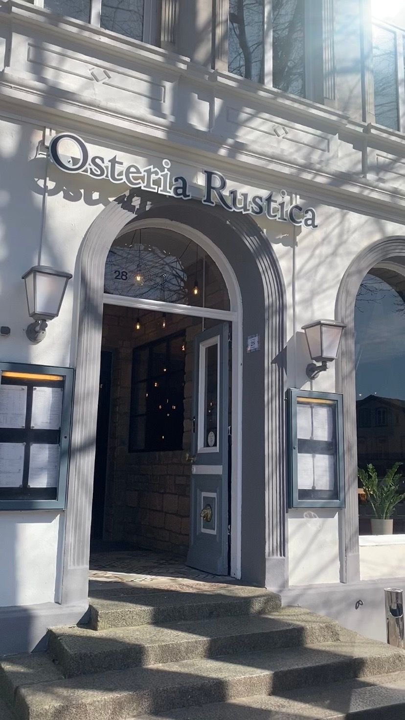 Eingang der Osteria Rustica mit offener Tür, Treppe und Laternen. Die Fassade ist weiß und das Schild zeigt den Restaurantnamen.